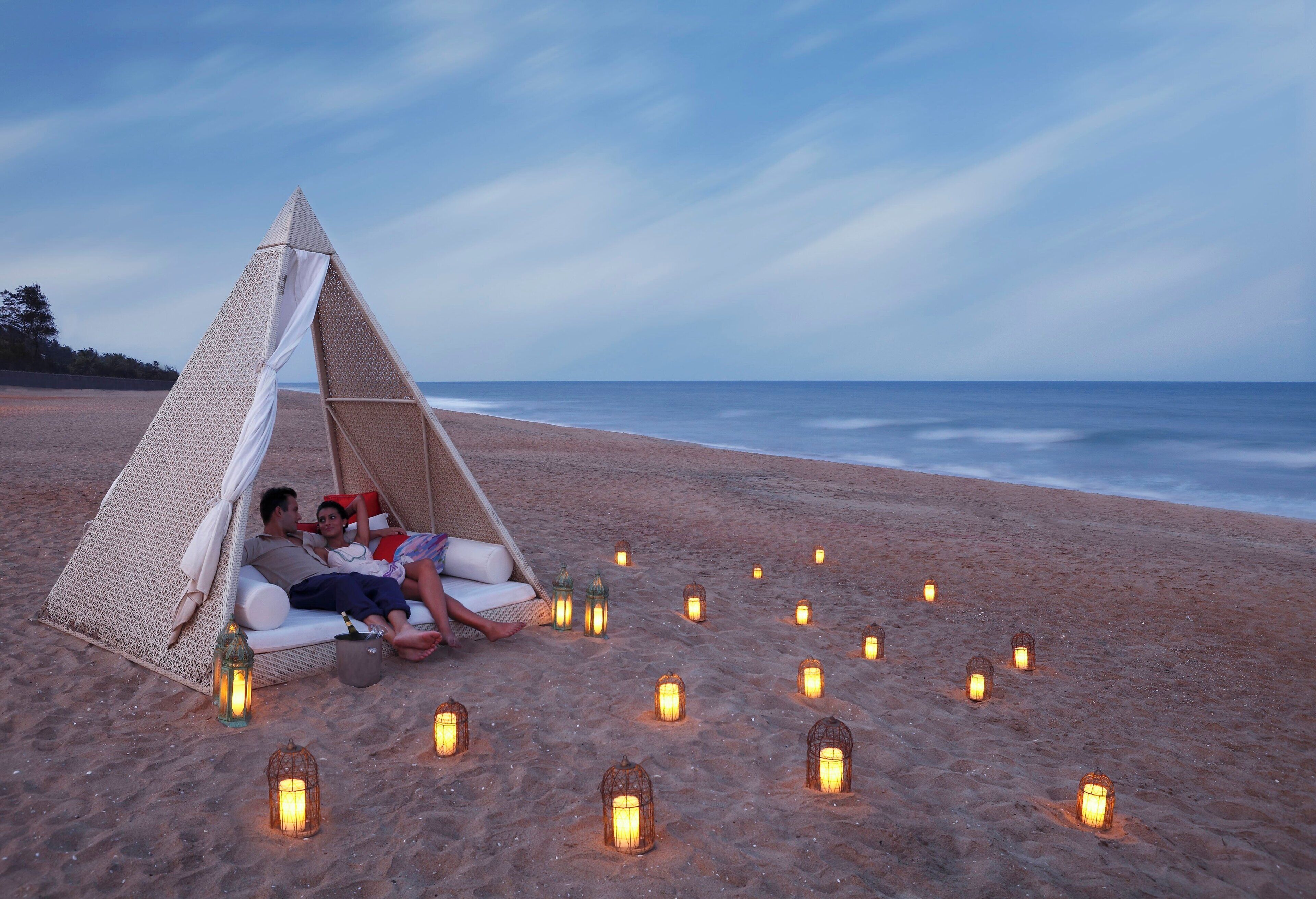 A beach scene with a couple relaxing on a white sofa under a triangular canopy, surrounded by sand and small lanterns.
