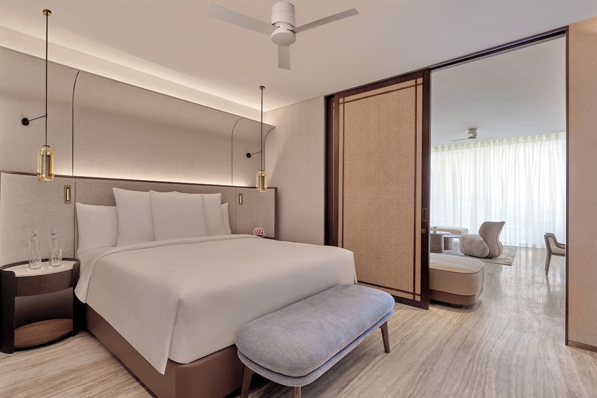 A modern hotel room with a large bed, bedside tables, a bench, a ceiling fan, and a view of a balcony with seating.