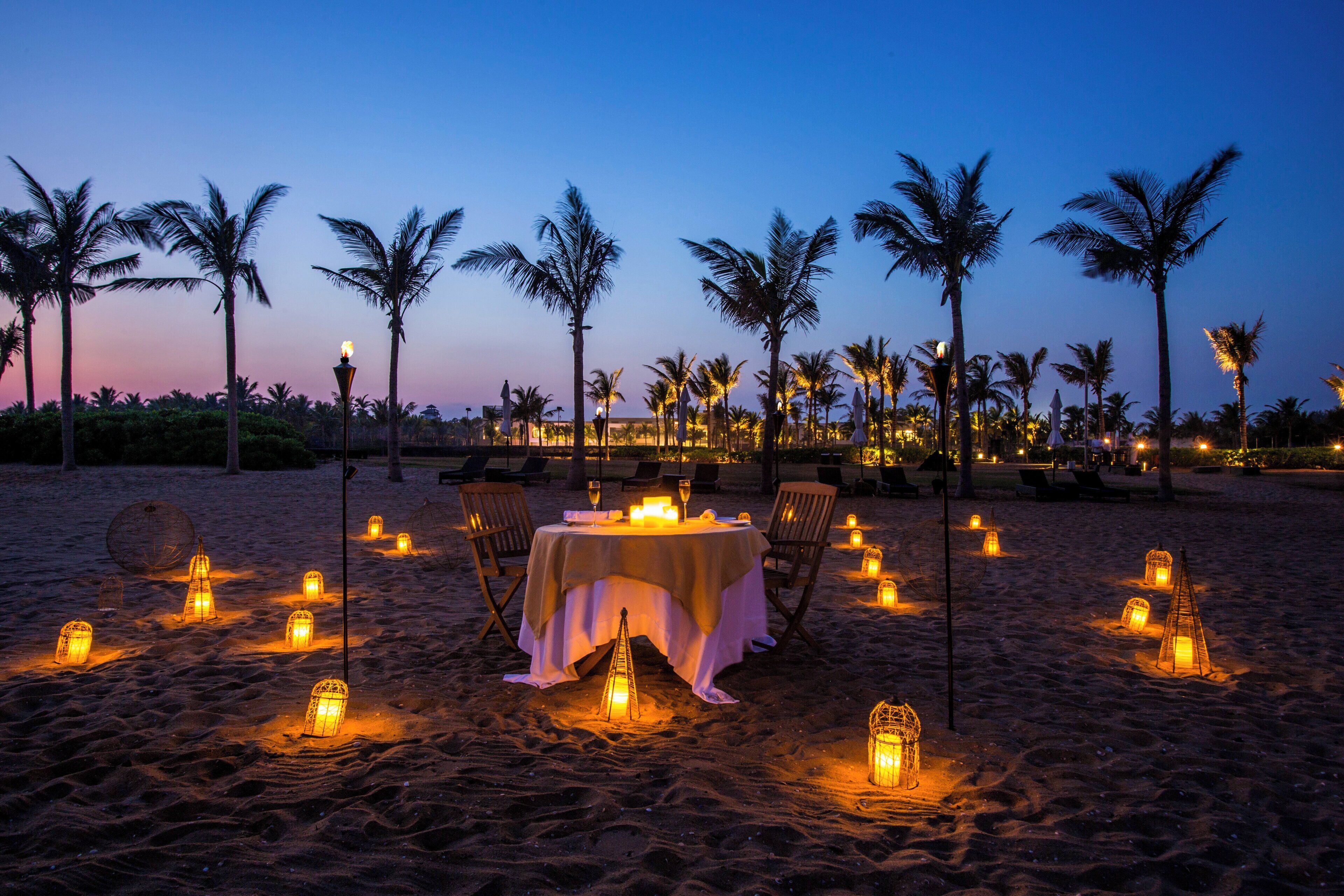 A beachside dining setup with a table and chairs, surrounded by palm trees and lanterns.