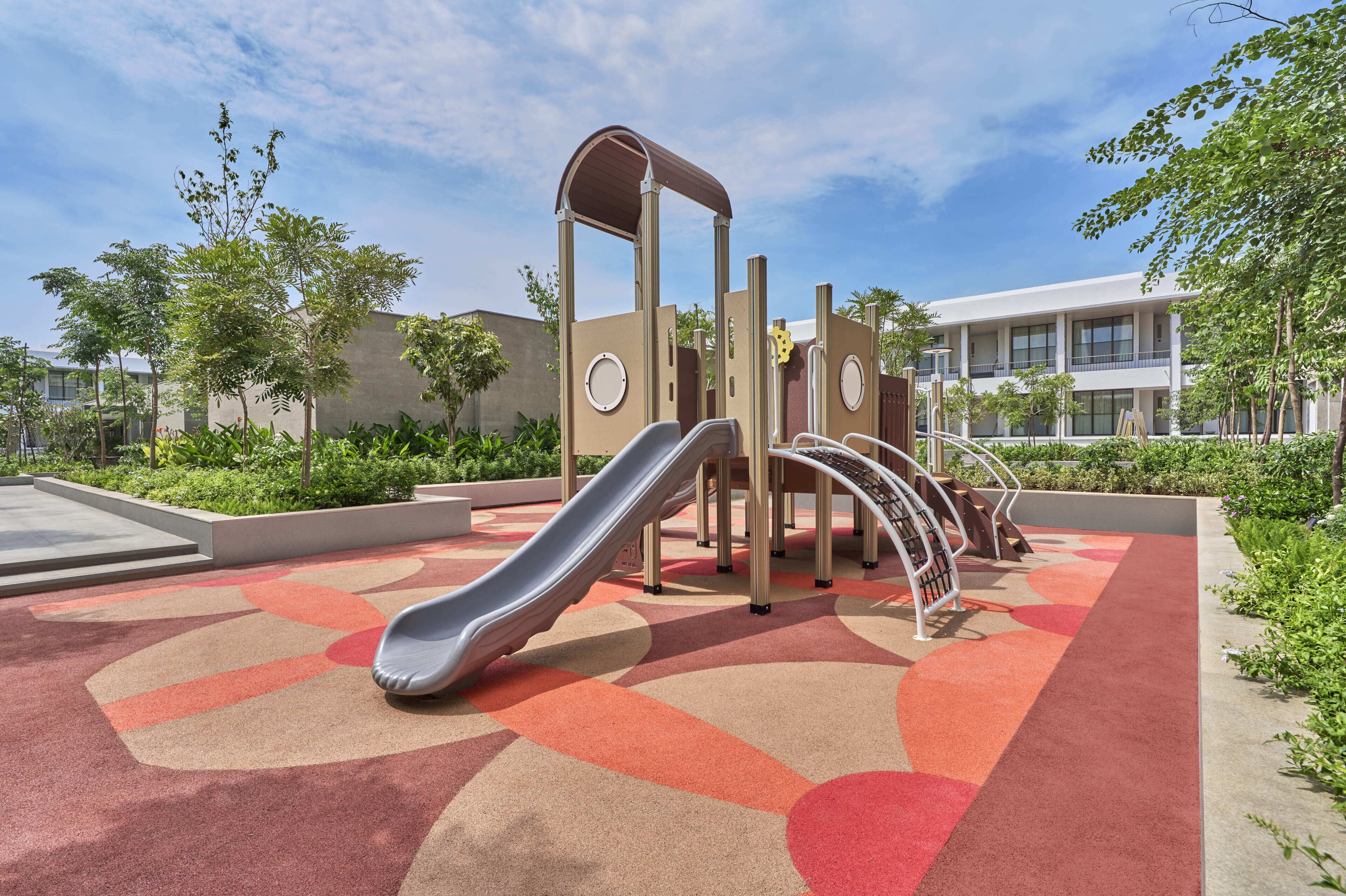 A playground with a slide and climbing structures, surrounded by greenery and a building in the background.