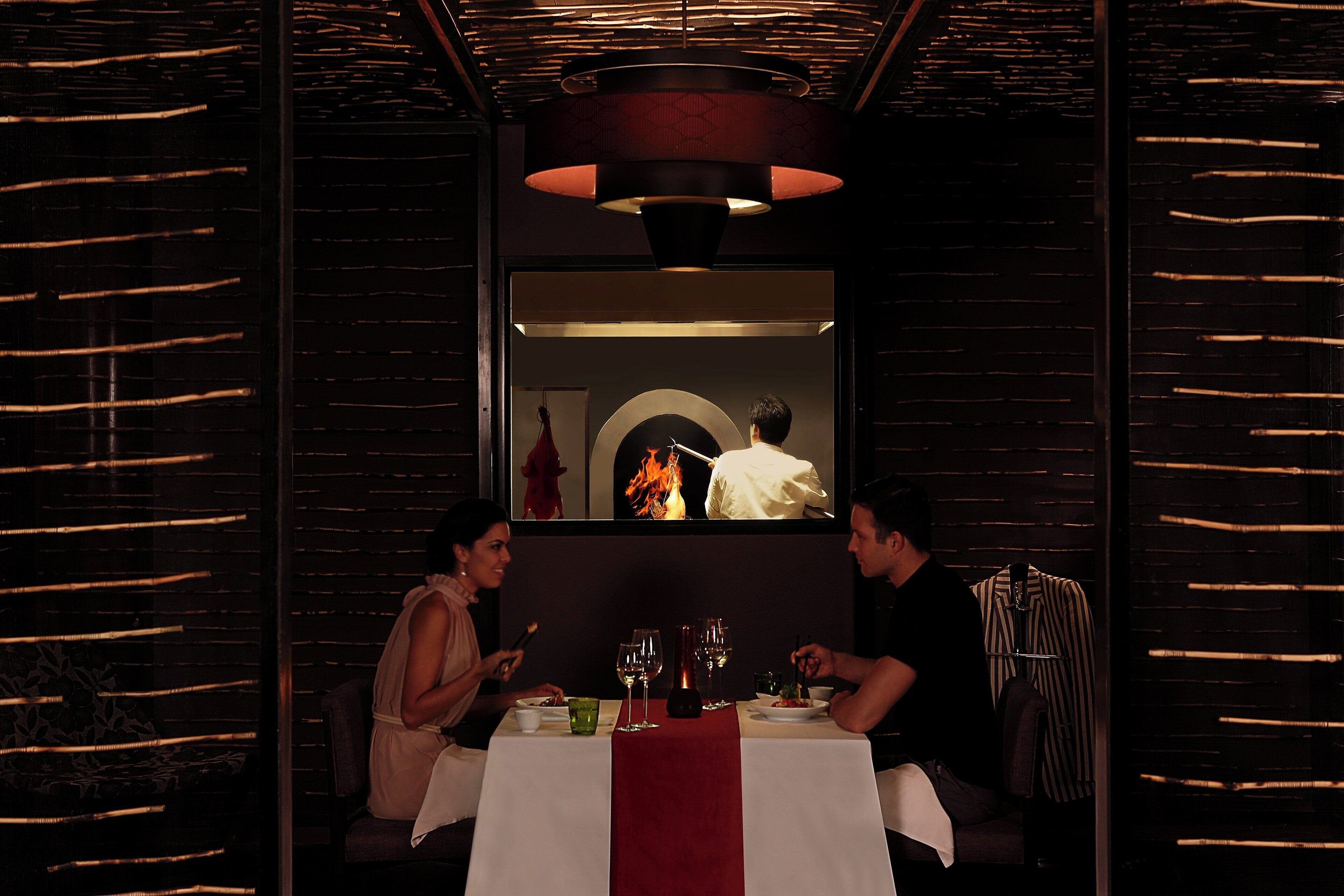 Restaurante com iluminação suave, mesa posta para dois, um jogo americano vermelho e um lustre pendurado acima.