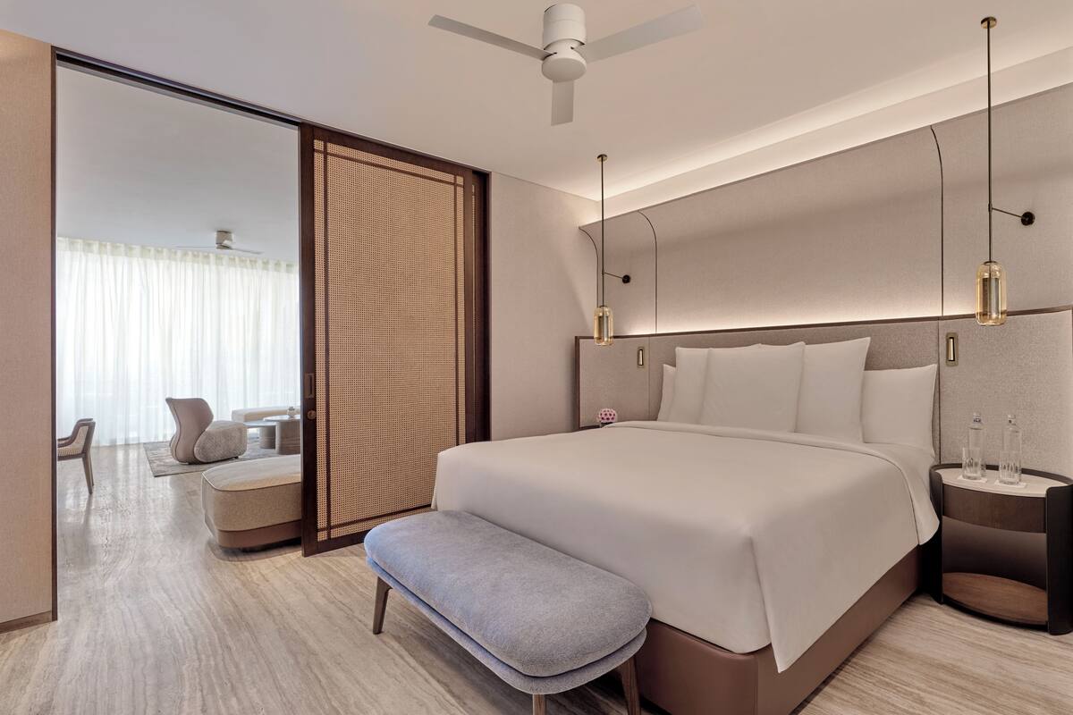 Ein modernes Hotelzimmer mit einem großen Bett, einer Bank, einem Nachttisch mit Lampe und Blick durch eine Schiebetür.