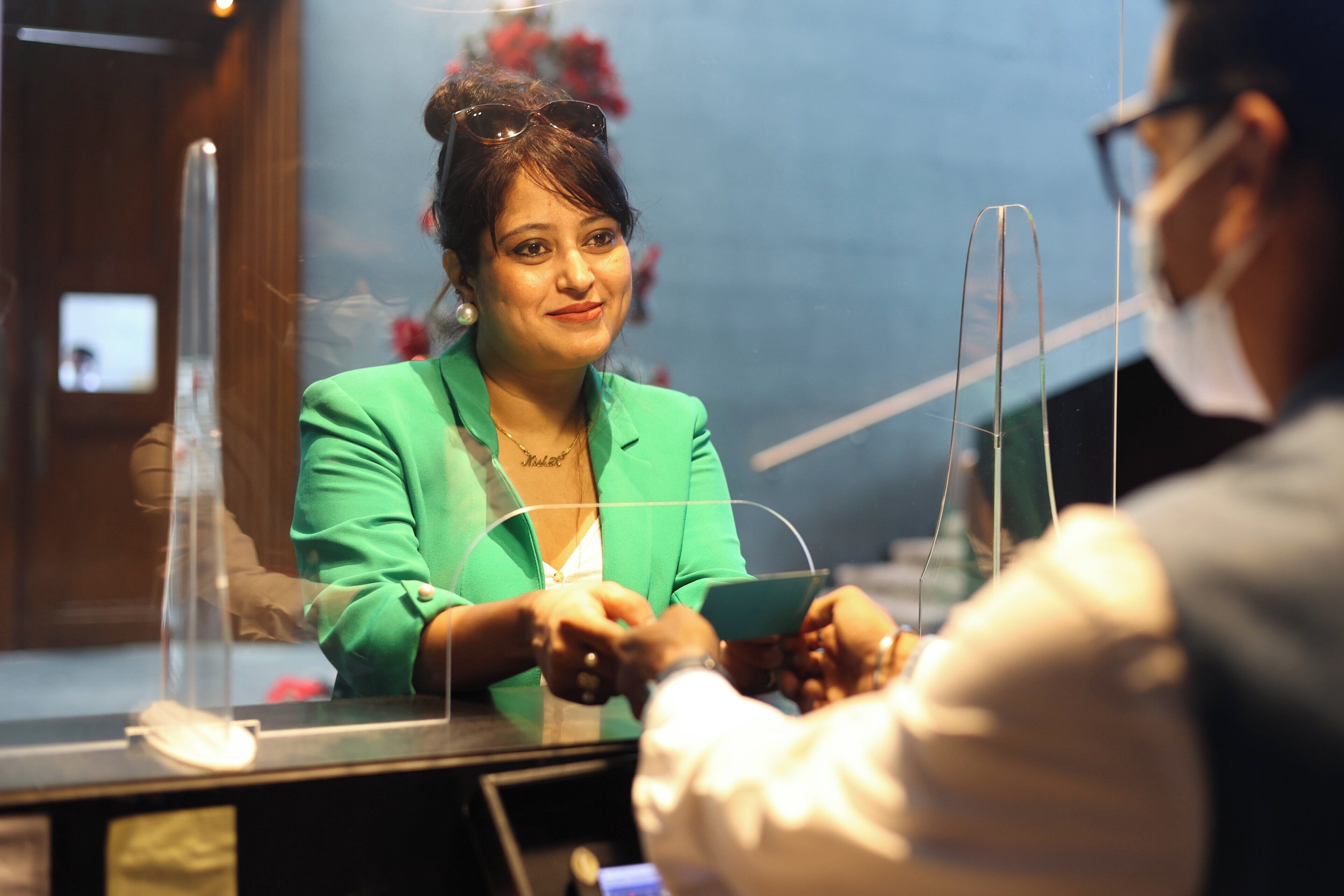 A person in a green blazer at a counter, possibly in a hotel or restaurant, with a clear plastic barrier between them.