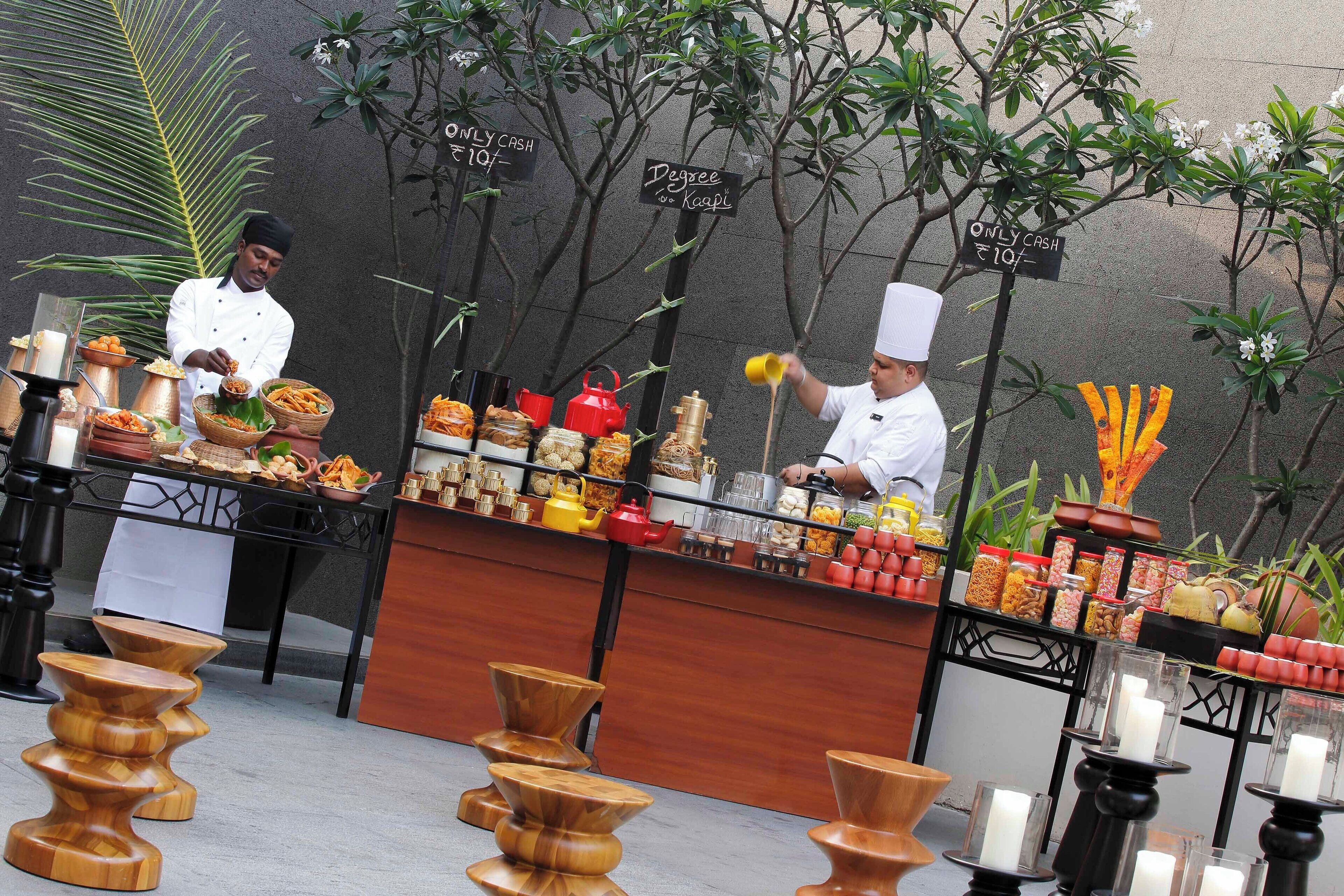 A food stall with various snacks and condiments, cash-only payment signs, and a backdrop of trees and a wall.