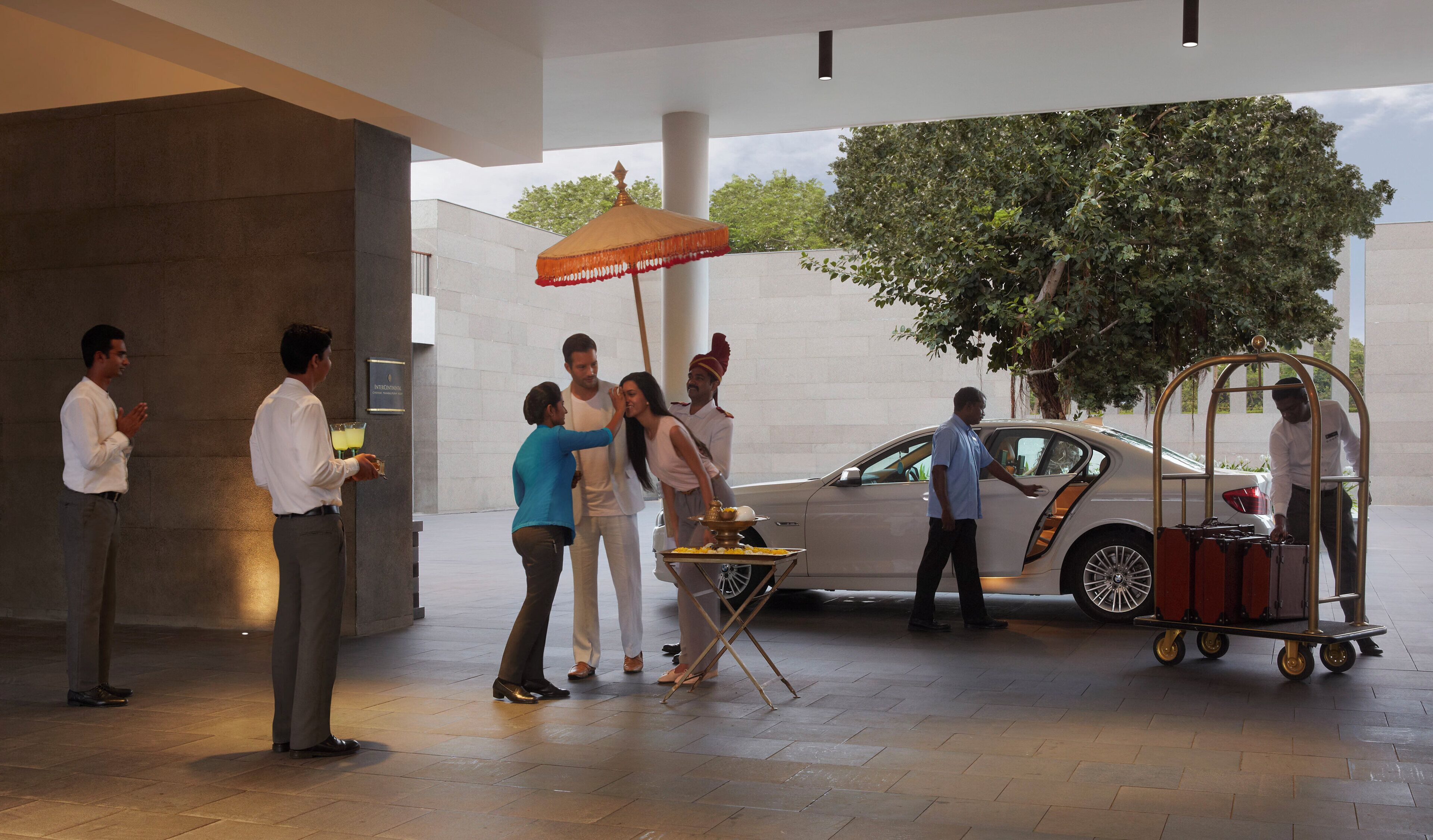 A group of people near a car with a service cart, a large umbrella, and a tree in the background.