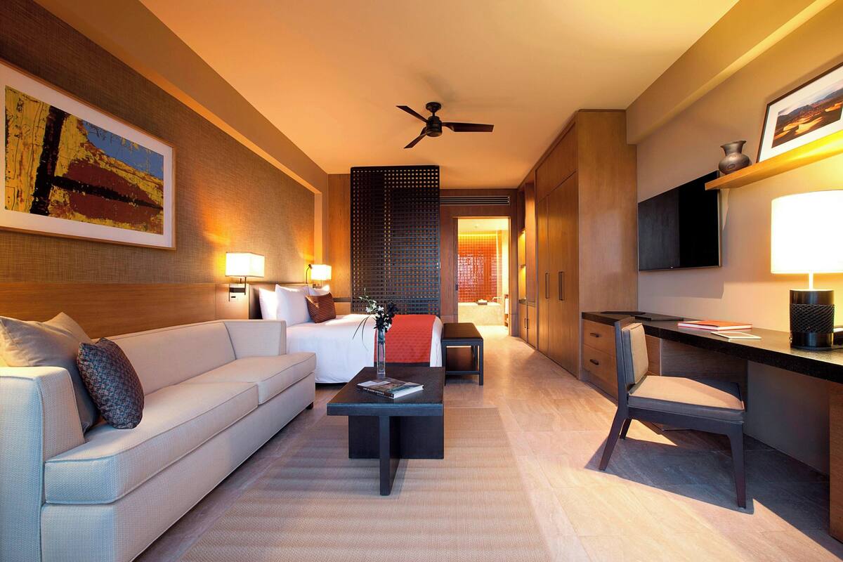 Een moderne hotelkamer met een bank, een bijzettafeltje, een bureau en een stoel. Er is een plafondventilator, een schilderij aan de muur en een lamp.