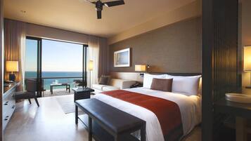 A hotel room with a large bed, a desk, a chair, a bench, a nightstand, and a view of the ocean.