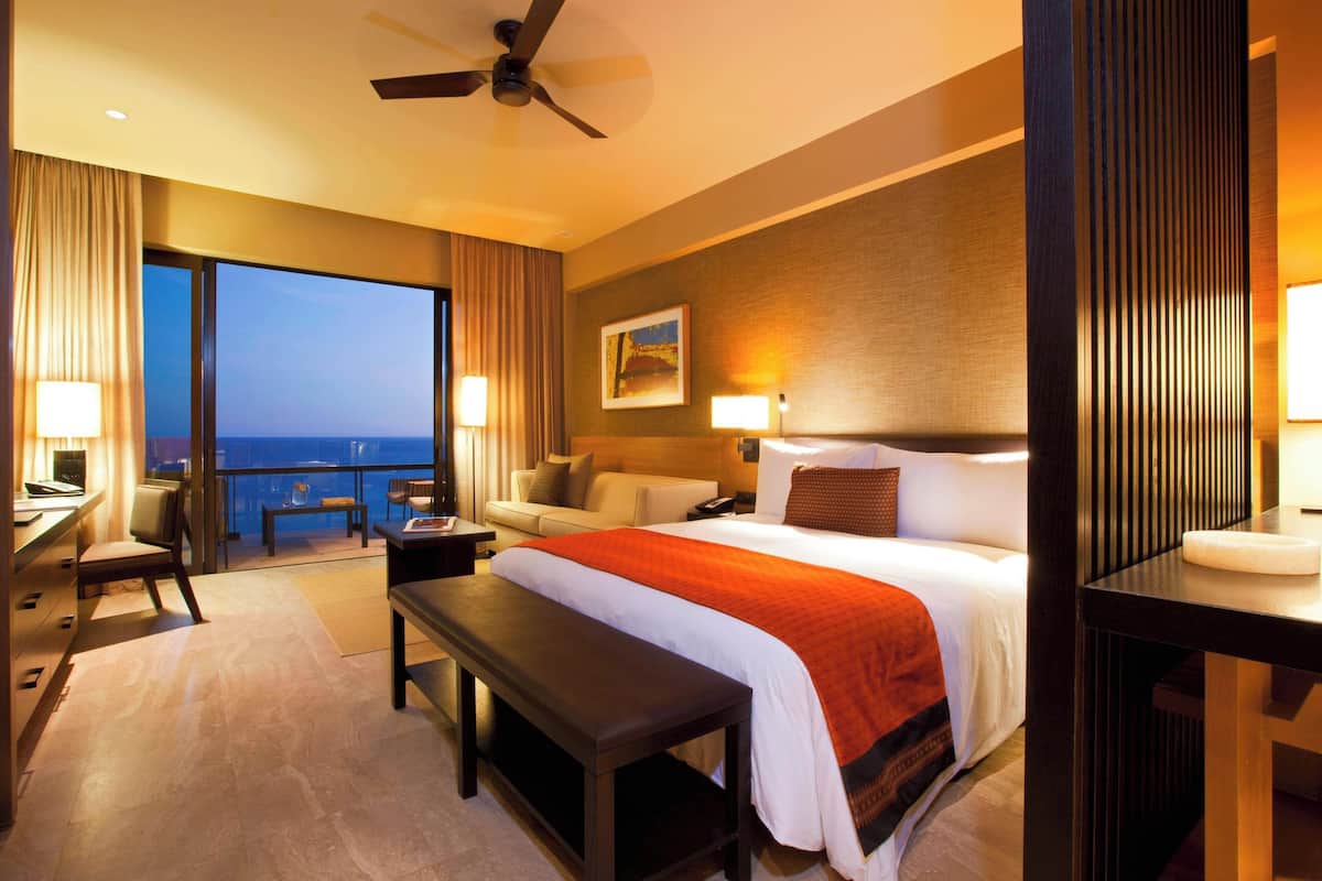 Een hotelkamer met een groot bed, een bureau, een stoel, een bankje, een bureaulamp, een nachtkastje en uitzicht op de oceaan.