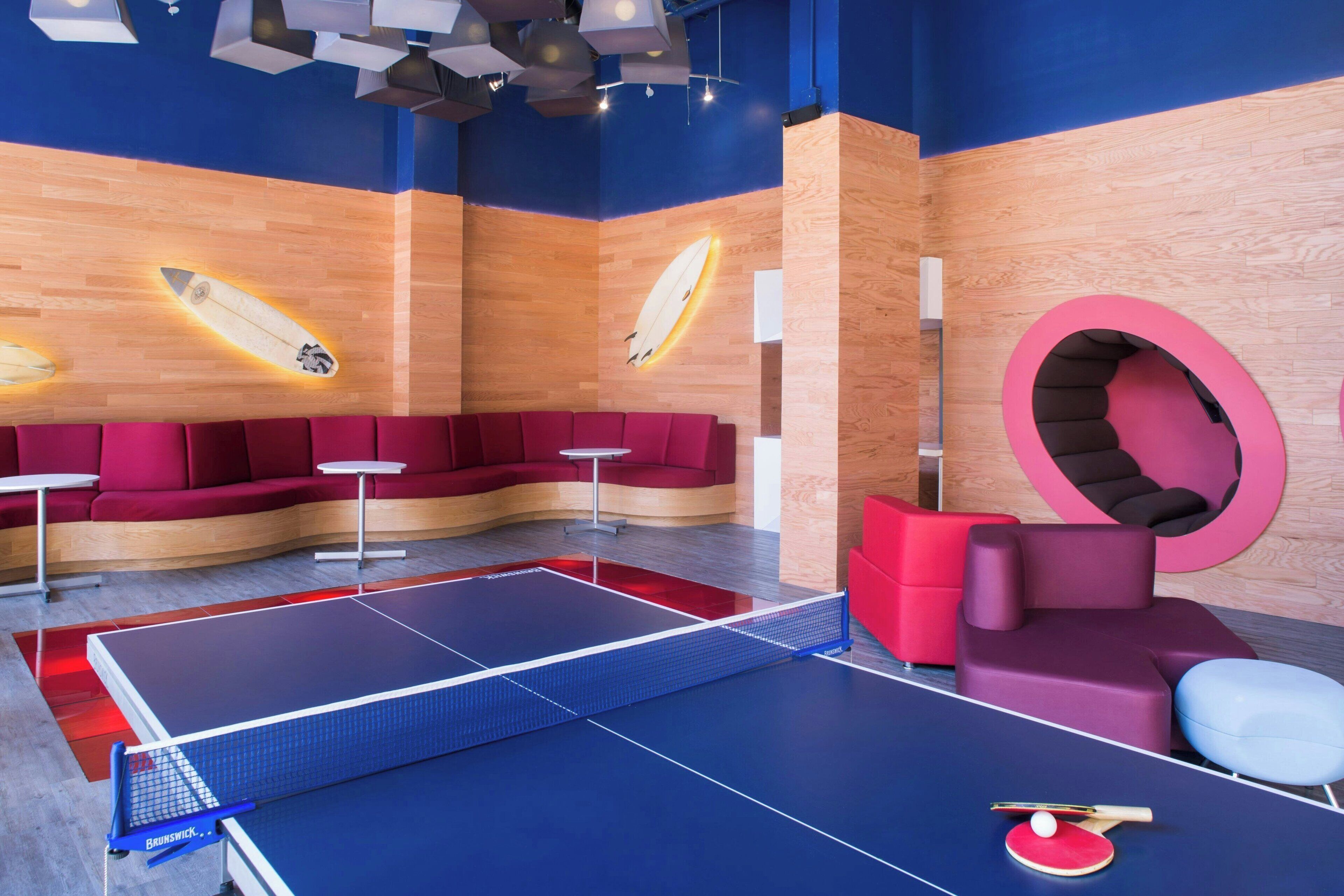 A ping pong table with a net, red and purple seating, and a surfboard mural on the wall.