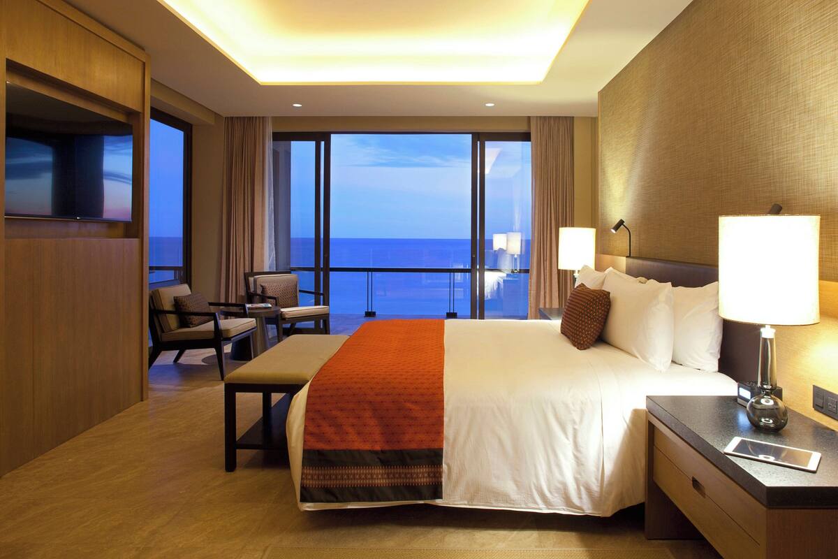 Een hotelkamer met een groot bed, een bureau met lamp, een stoel en uitzicht op de oceaan.