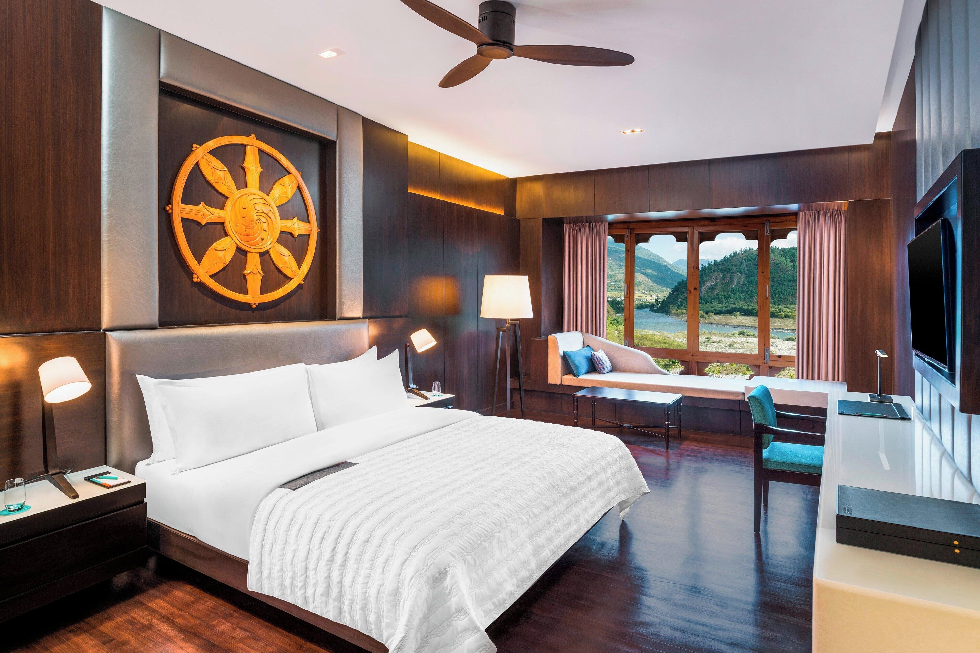 Habitación de hotel moderna con una cama grande, un televisor montado en la pared, un ventilador de techo y vistas a un paisaje montañoso a través de una ventana.