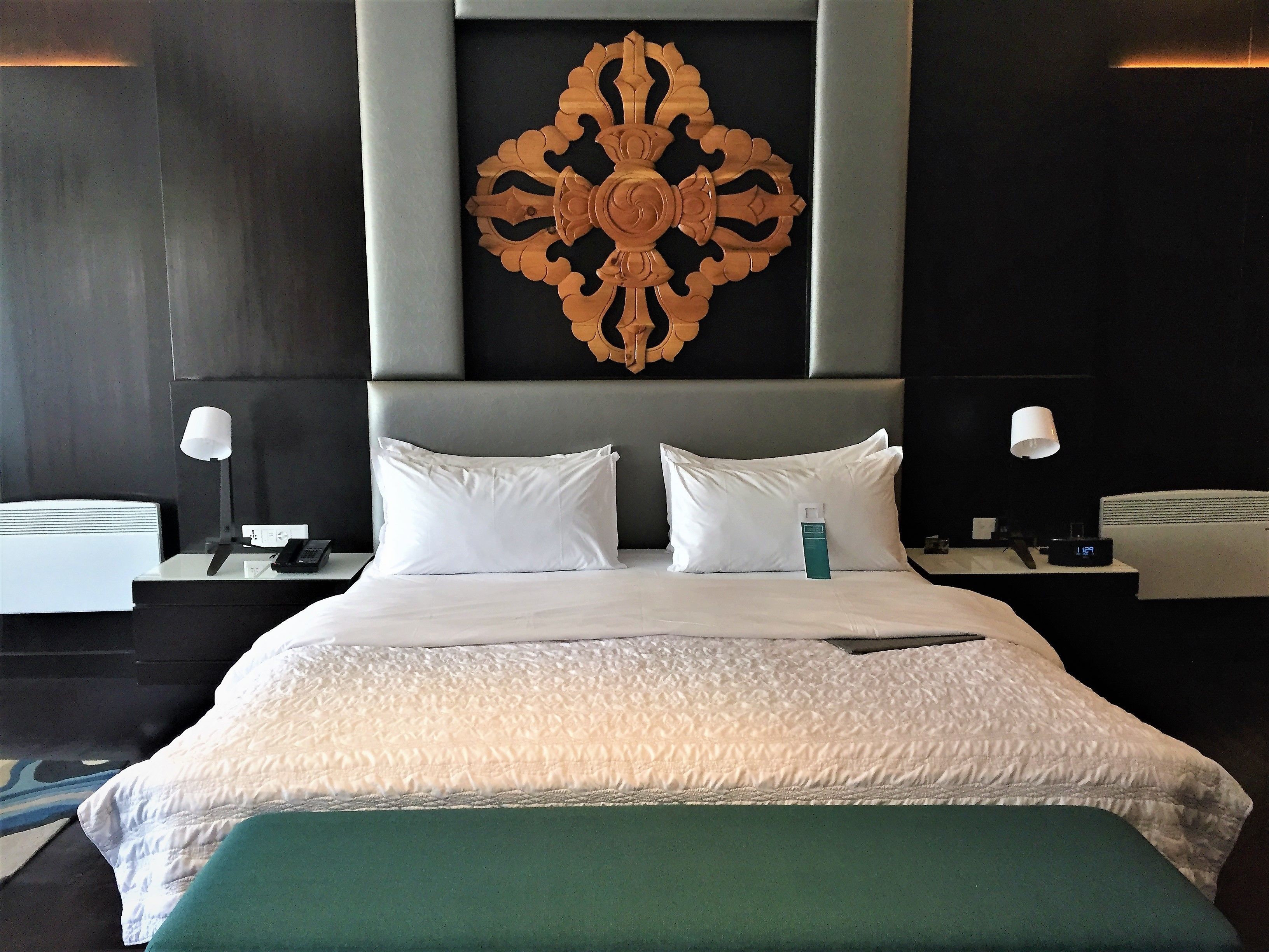 A hotel room with a large bed, two bedside tables, a nightstand, a decorative wall piece, and a green accent pillow.