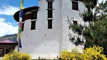 A traditional building with a white exterior and multiple windows, surrounded by yellow flowering bushes and a flagpole with colorful flags.