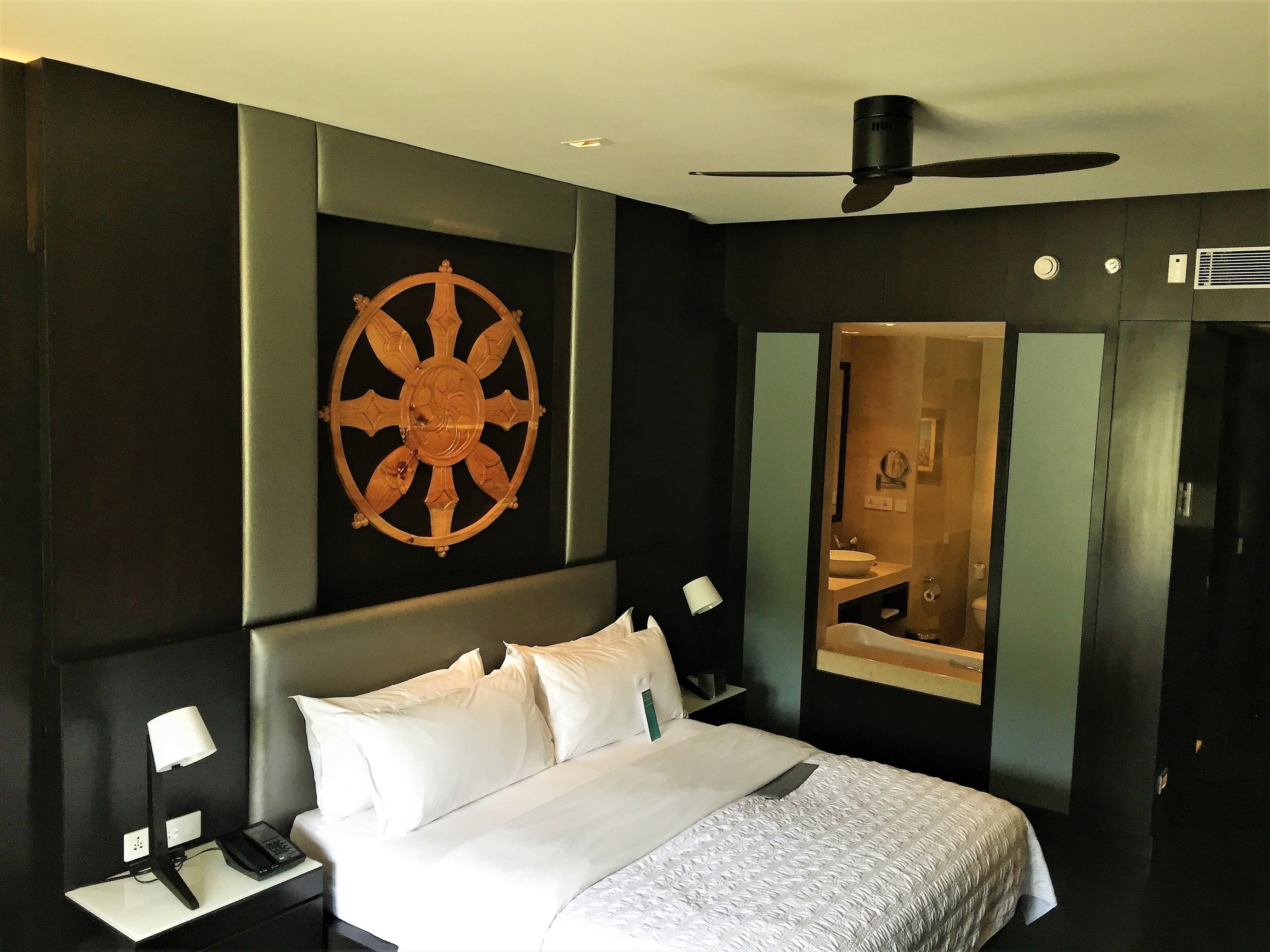 A hotel room with a bed, bedside tables, a wall-mounted ship's wheel, a ceiling fan, and a mirror.