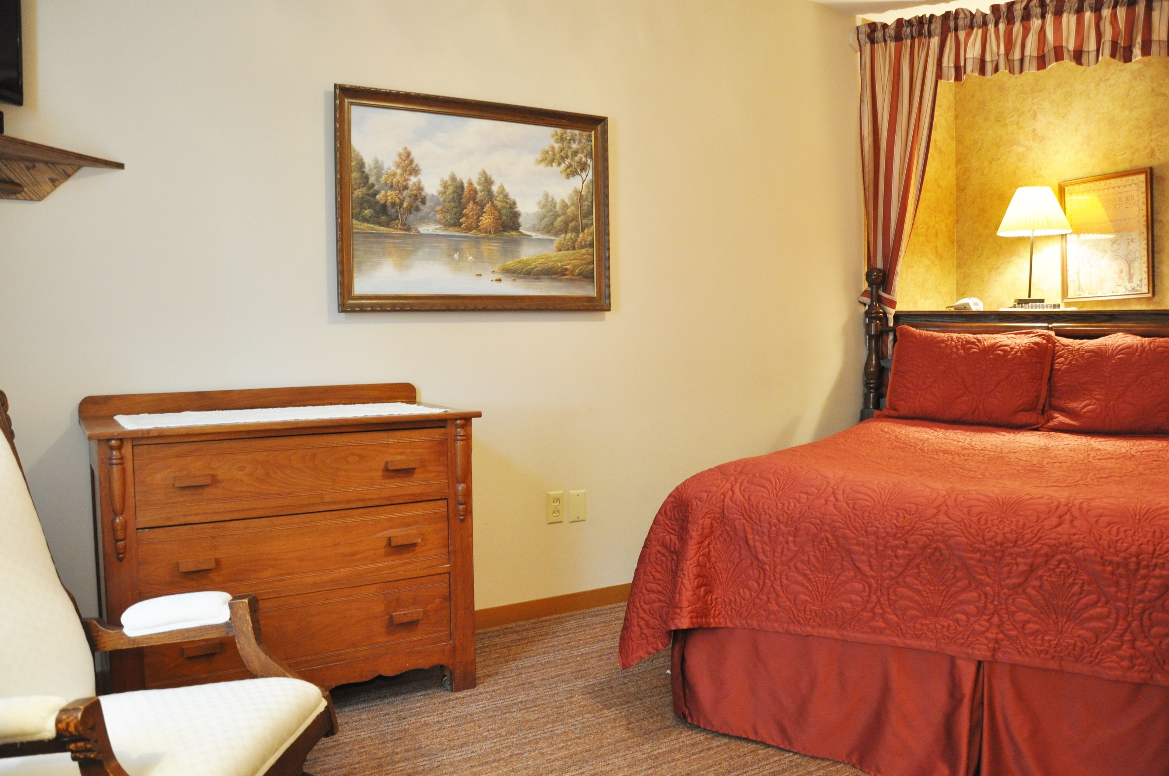 Une chambre d’hôtel avec une commode en bois, un lit avec une literie rouge, un tableau représentant un lac et une lampe sur la table de chevet.