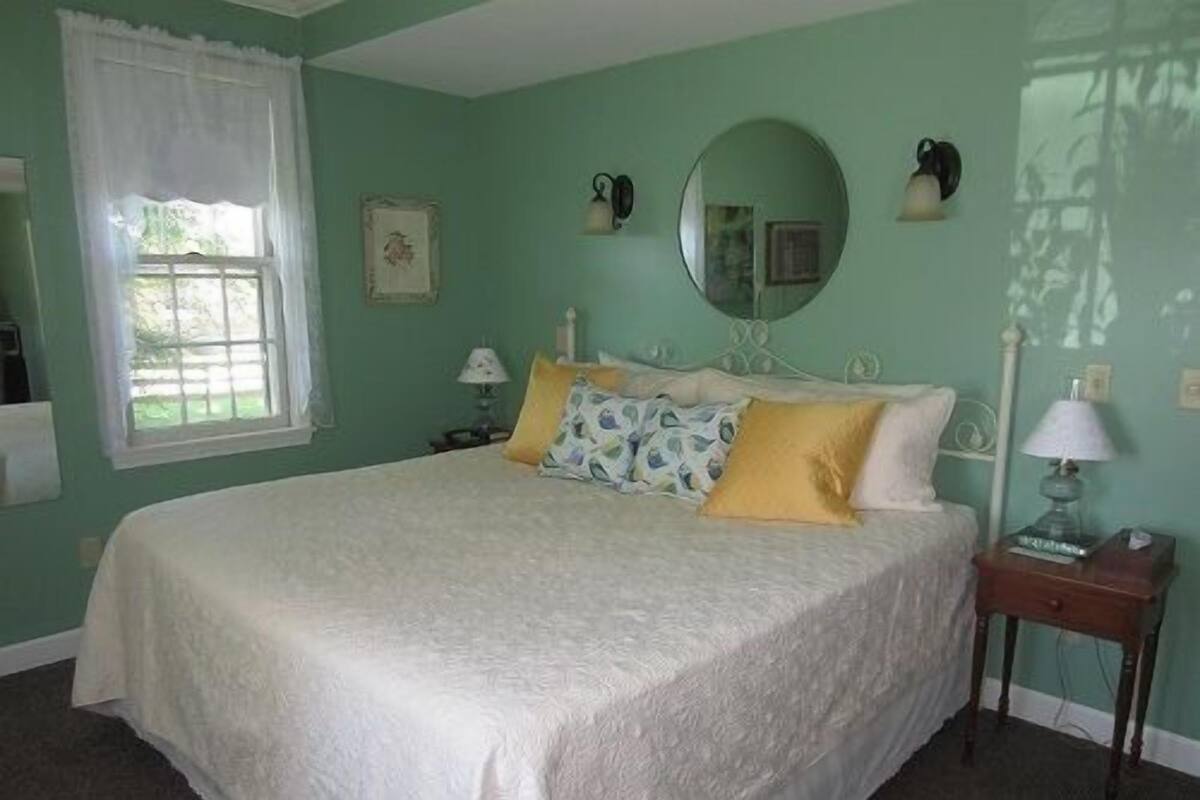 A bedroom with a large bed, a bedside table, a nightstand, a window with curtains, and a wall-mounted mirror.