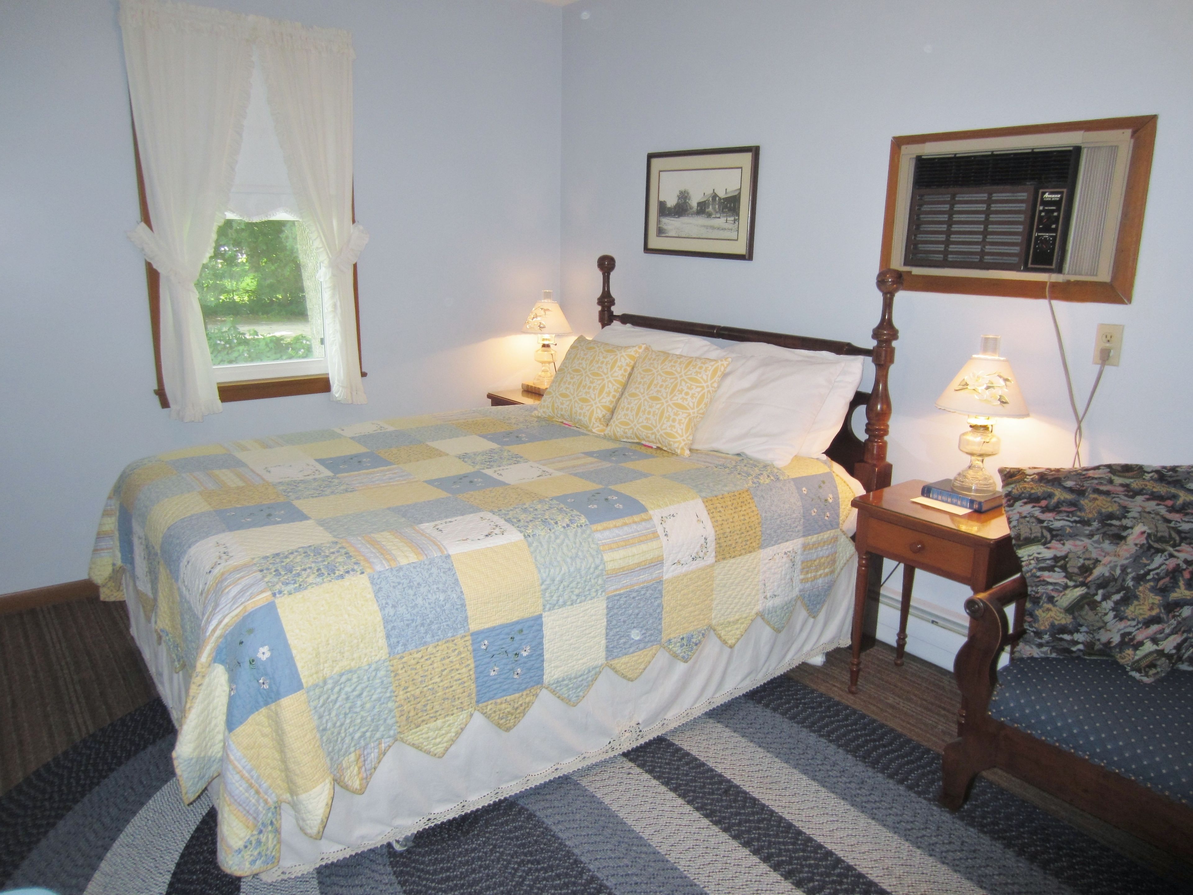 A bedroom with a bed, a nightstand, a lamp, a window with curtains, a wall-mounted air conditioner, and a framed picture.