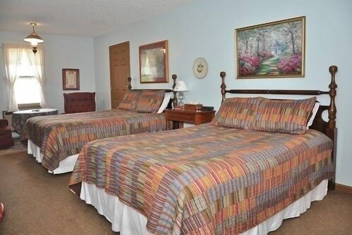 A hotel room with two beds, a small table, a chair, a painting on the wall, and a window with curtains.