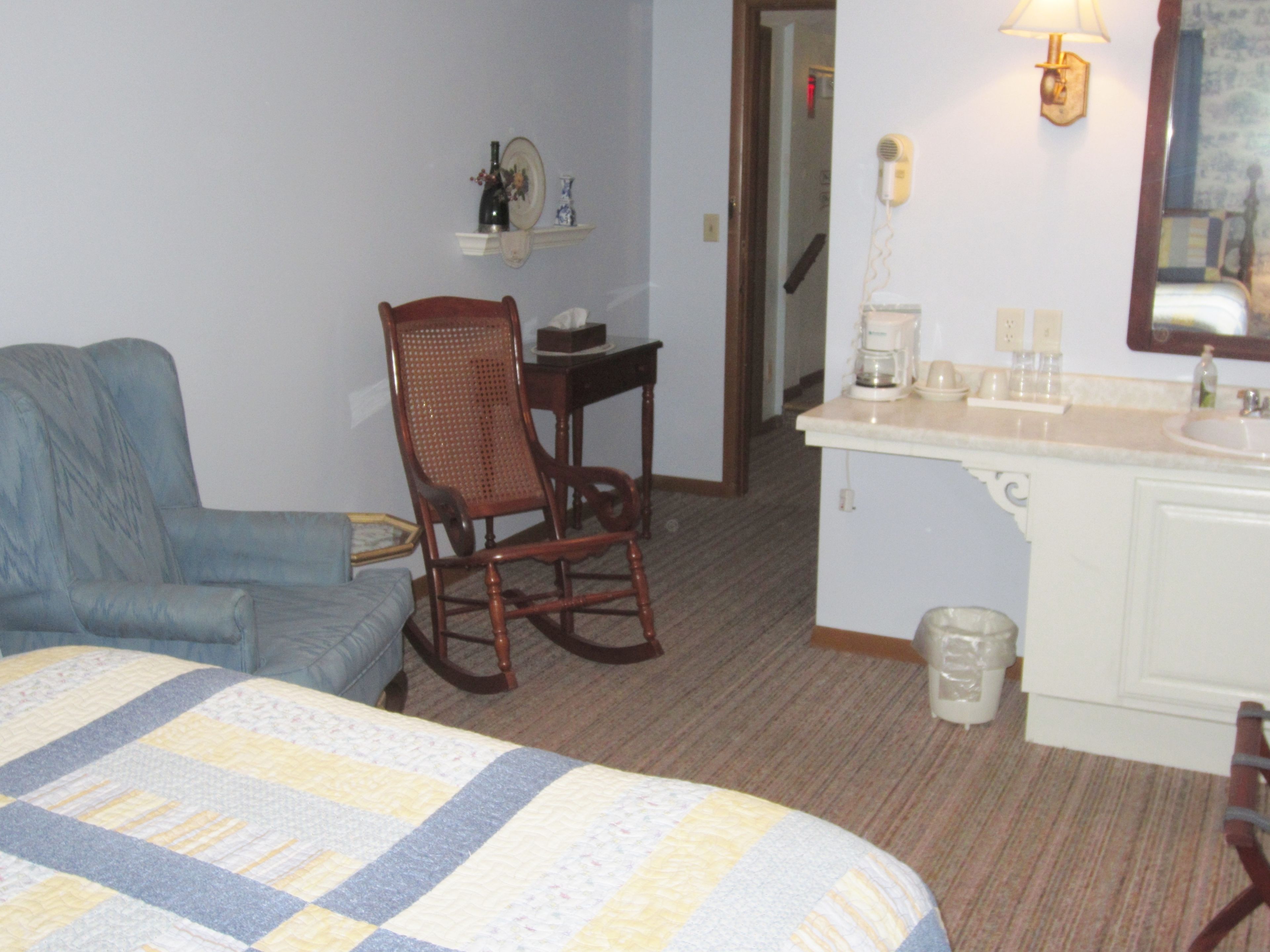 A hotel room with a bed, a rocking chair, a desk, a small shelf with a bottle and a plate, a coffee maker, and a mirror.