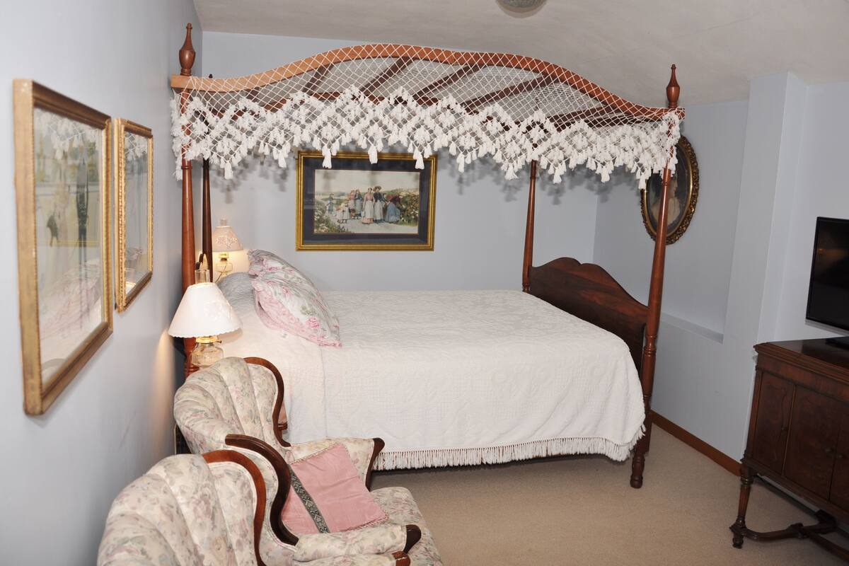 A bedroom with a four-poster bed, a chair, a lamp, a television, and framed pictures on the wall.