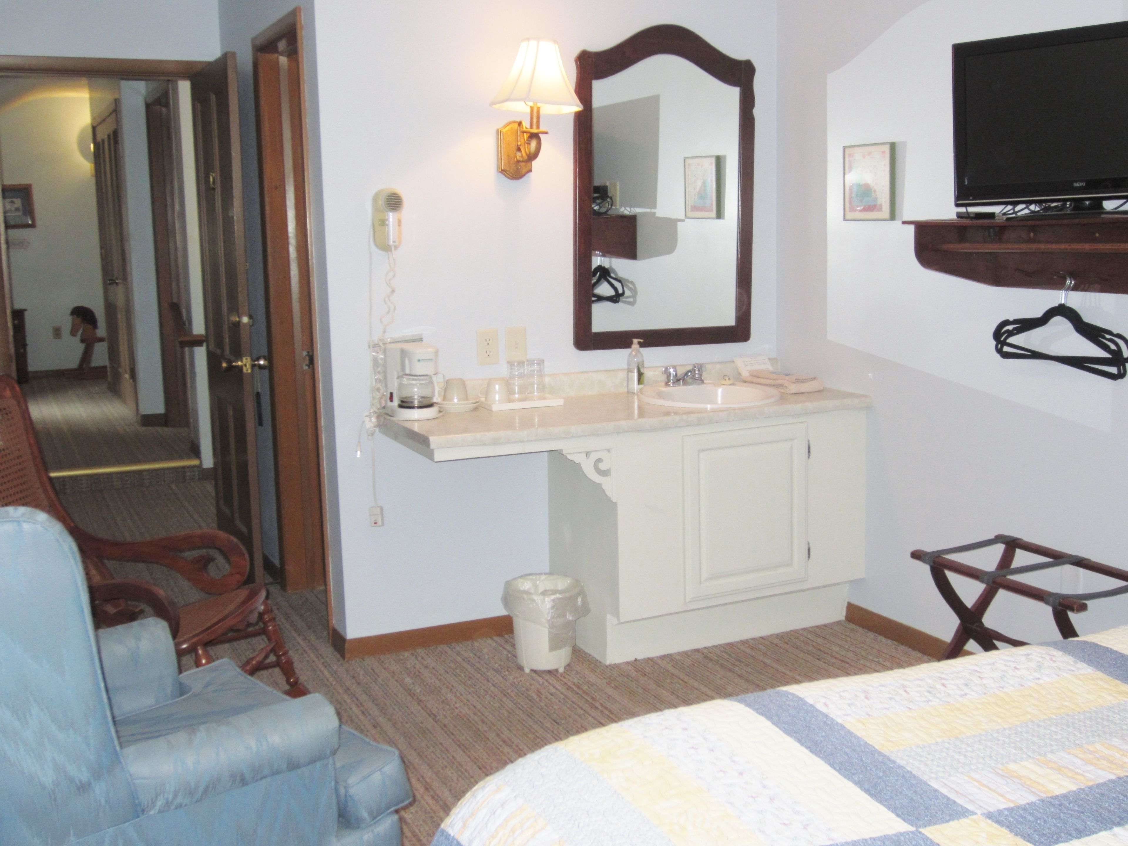 A hotel room with a bed, a desk, a chair, a mirror, a television, a sink, and a trash can.