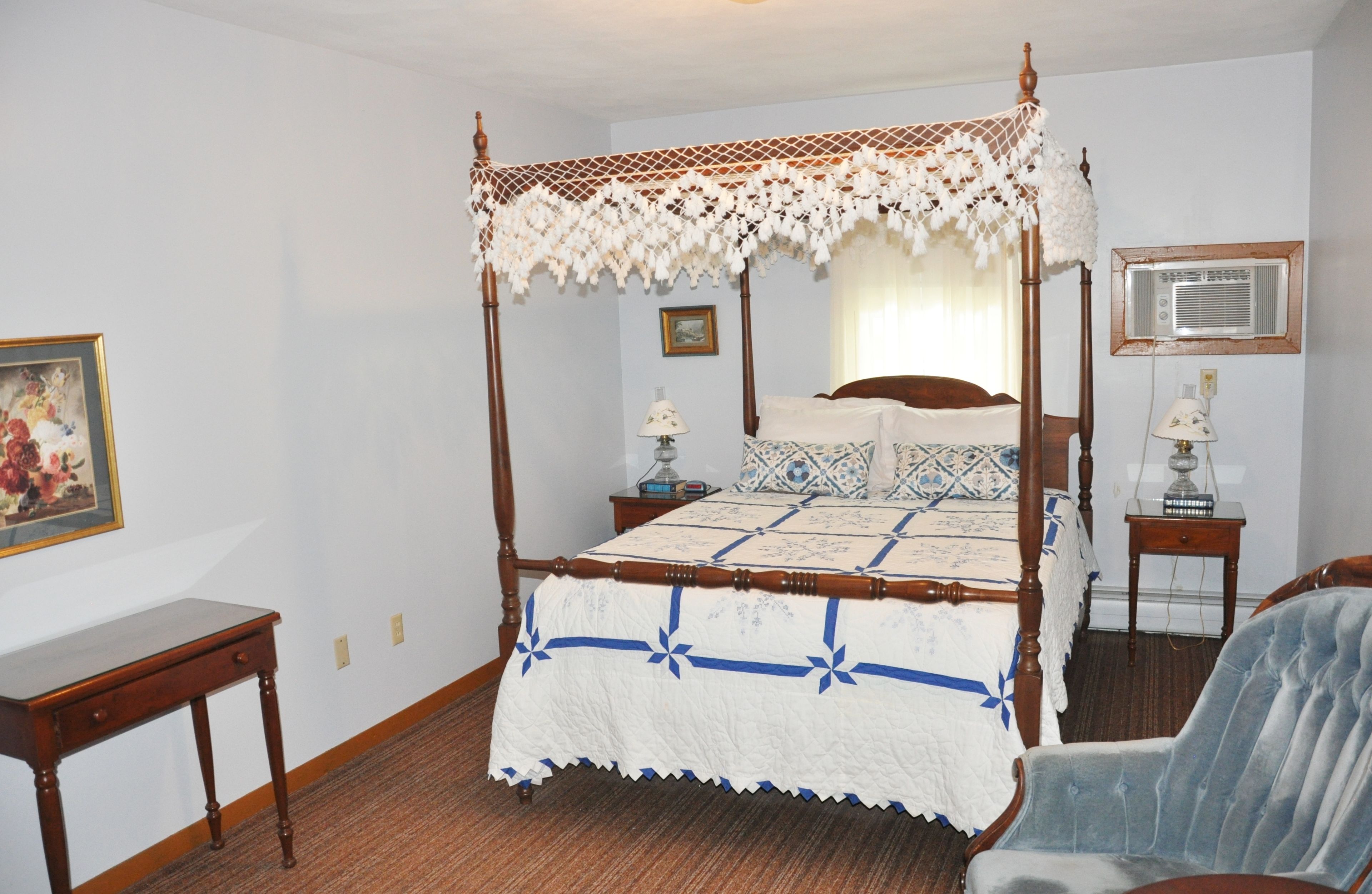 A four-poster bed with a white and blue quilt, wooden nightstands, a desk, and a chair.