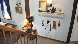 A room with a wooden staircase, a table with a lamp, a welcome sign, and wall-mounted decorations including a framed picture, a shelf with a teapot, and various tools. - Die Heimat Country Inn (Homestead)