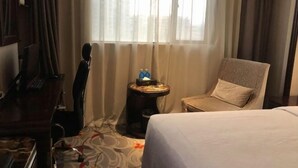 A hotel room with a bed, a desk, a chair, a small table with a bottle and a tissue box, and a window with curtains.