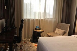 Ein Hotelzimmer mit einem Bett, einem Schreibtisch, einem Stuhl, einem kleinen Tisch mit einer Flasche und einer Taschentuchbox sowie einem Fenster mit Vorhängen.