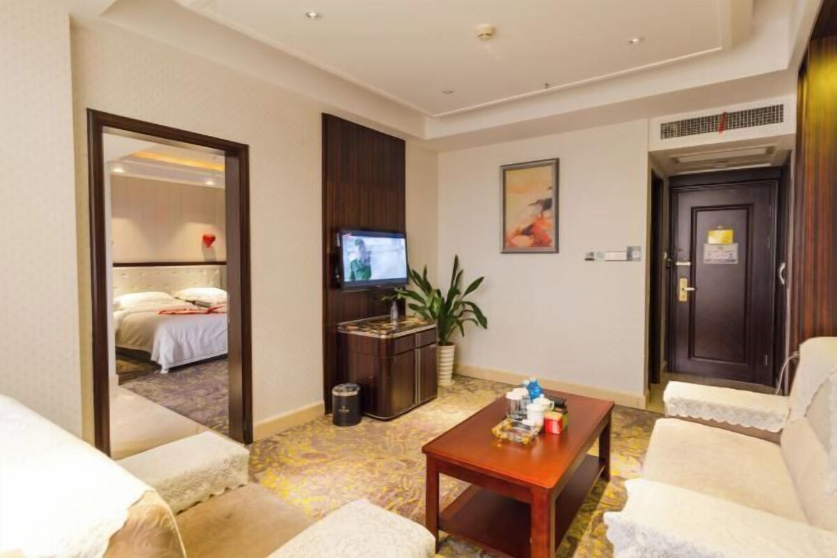 Una habitación de hotel con una cama, televisión, una mesita con objetos, una silla y una puerta.