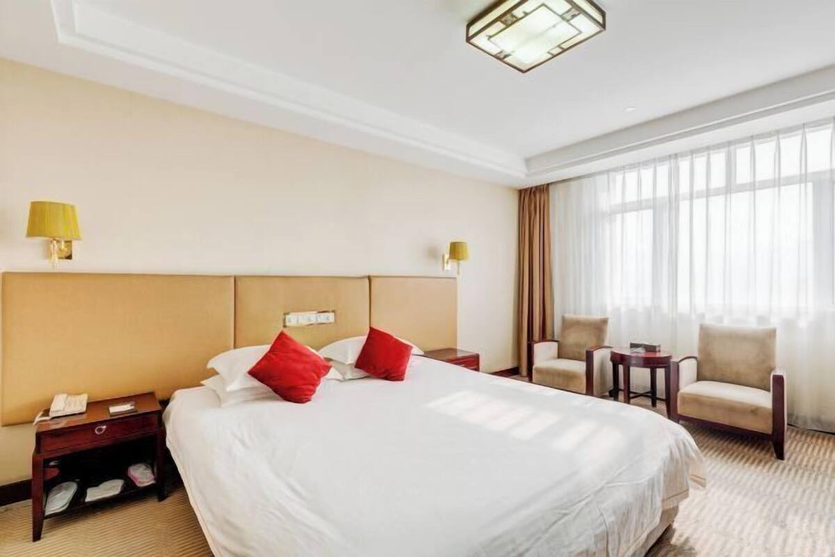 Una habitación de hotel con una cama grande, dos mesitas de noche, una silla, una pequeña mesa y un ventanal con cortinas.