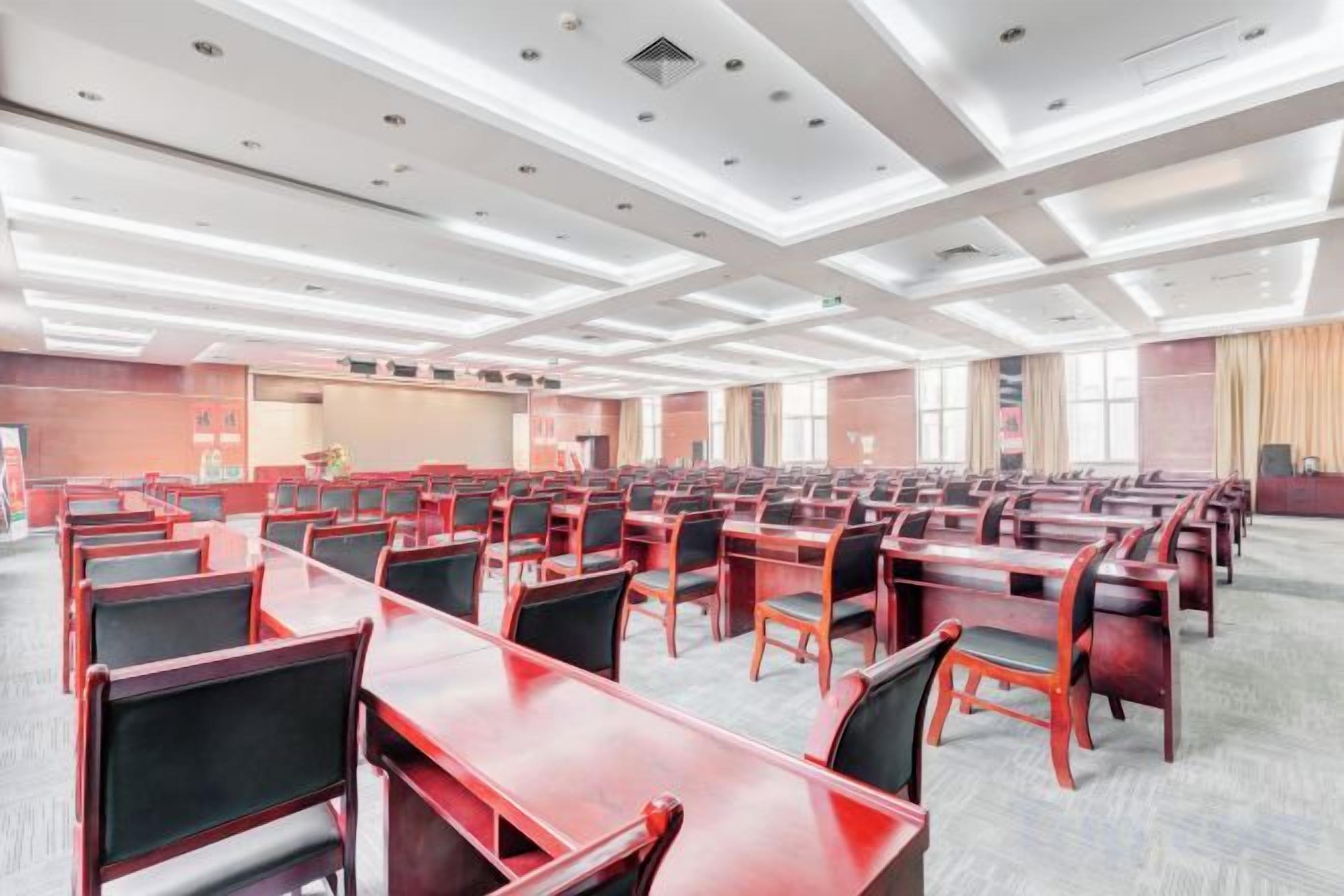 Une salle de conférence avec des rangées de tables et de chaises, une scène et de grandes fenêtres.