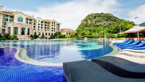 A large swimming pool with a curved edge, surrounded by lounge chairs and a building with multiple balconies. - Country Garden Sun City Phoenix Hotel (Shaoguan)