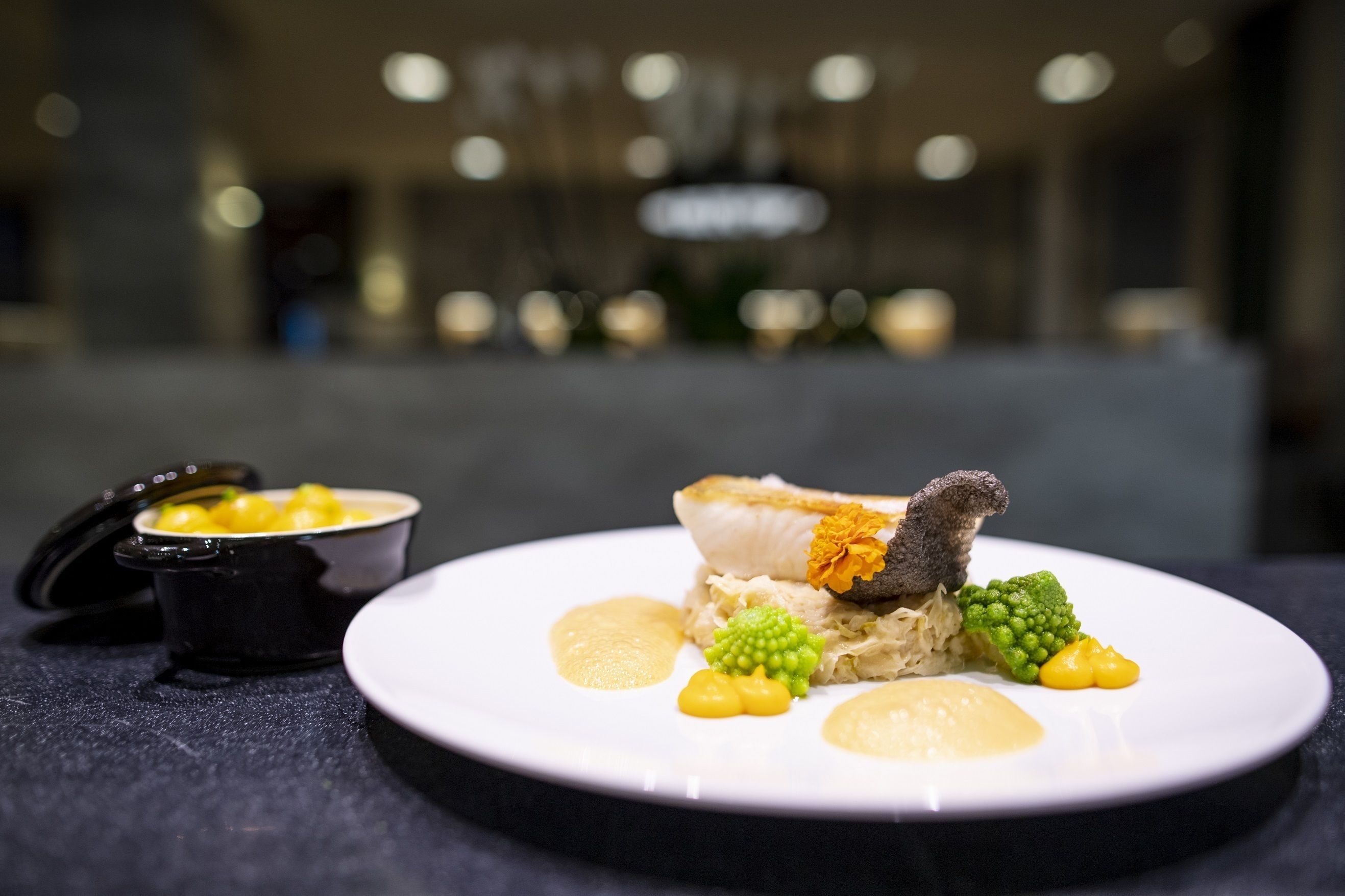 A plate of food with a fish, broccoli, and a small bowl of sauce.