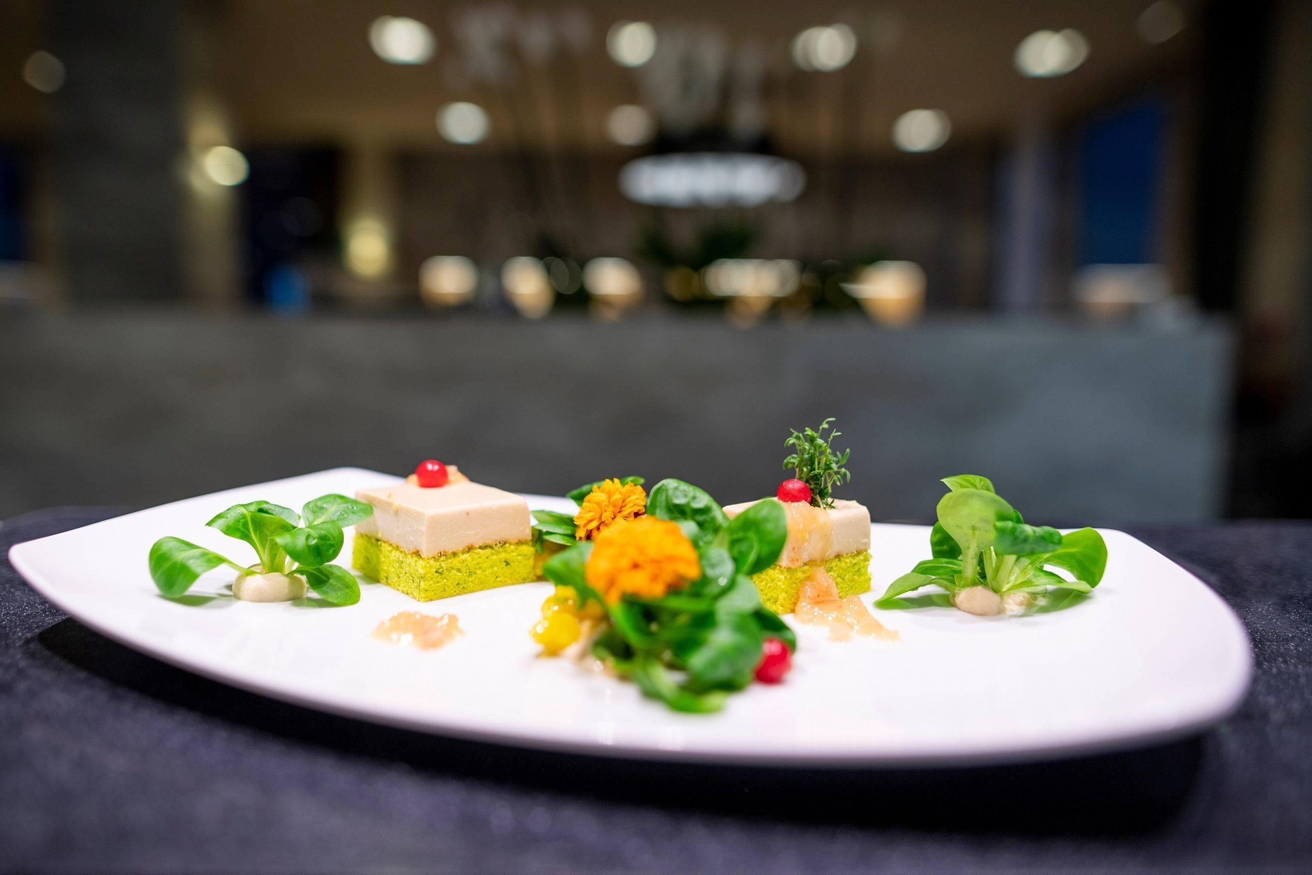 A white plate with three square desserts, each garnished with a cherry, green leaves, and a small sprig of herb.