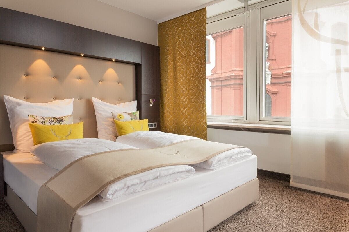 A hotel room with a large bed, a headboard with lights, a window with curtains, and a view of buildings outside.