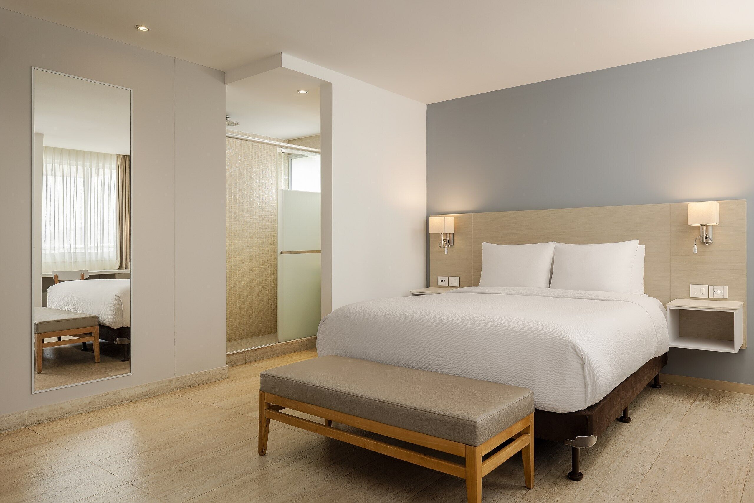 Habitació d'hotel moderna amb un llit gran, taules de nit, una banca, un mirall i una zona de dutxa.