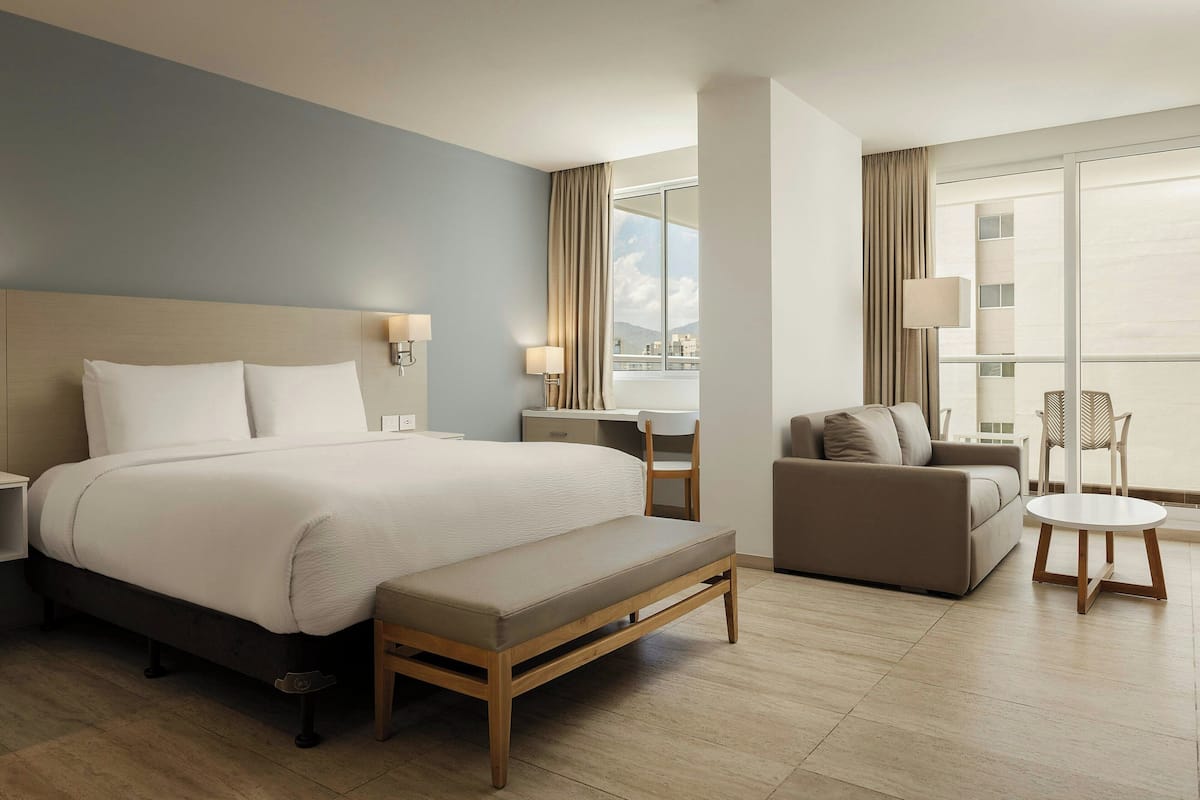 A modern hotel room with a large bed, a desk, a sofa, and a view of the city.