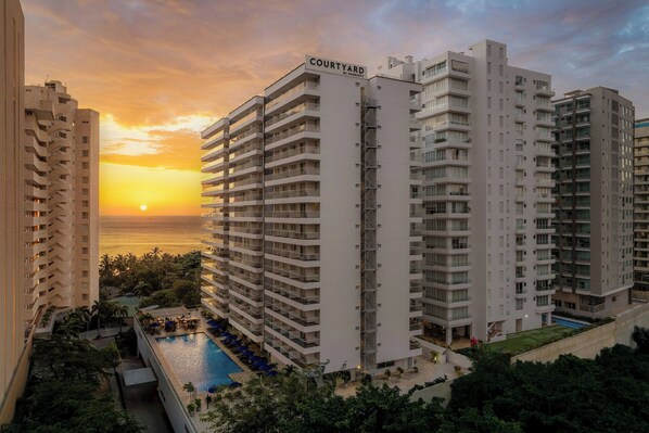 Um hotel moderno com várias varandas, uma piscina e vista para o pôr do sol sobre o oceano.