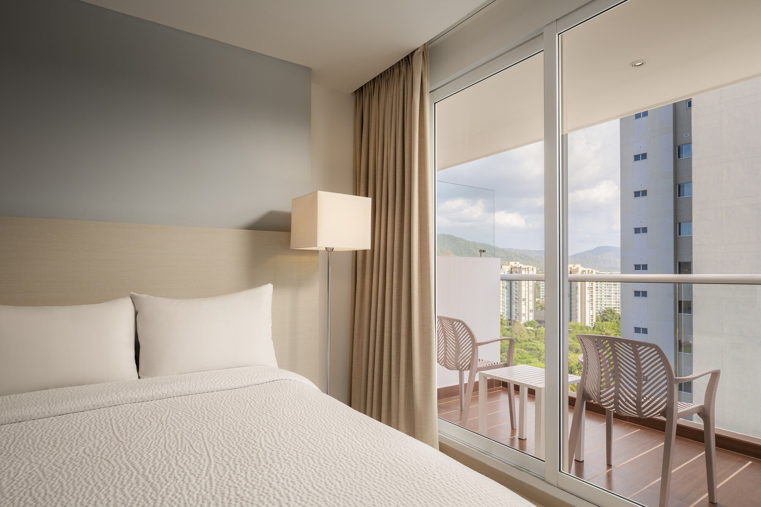 A hotel room with a large bed, a window with a view of buildings, and a balcony with chairs.