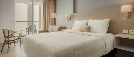 A hotel room with a large bed, a desk with a chair, a window with curtains, and wall-mounted lamps.