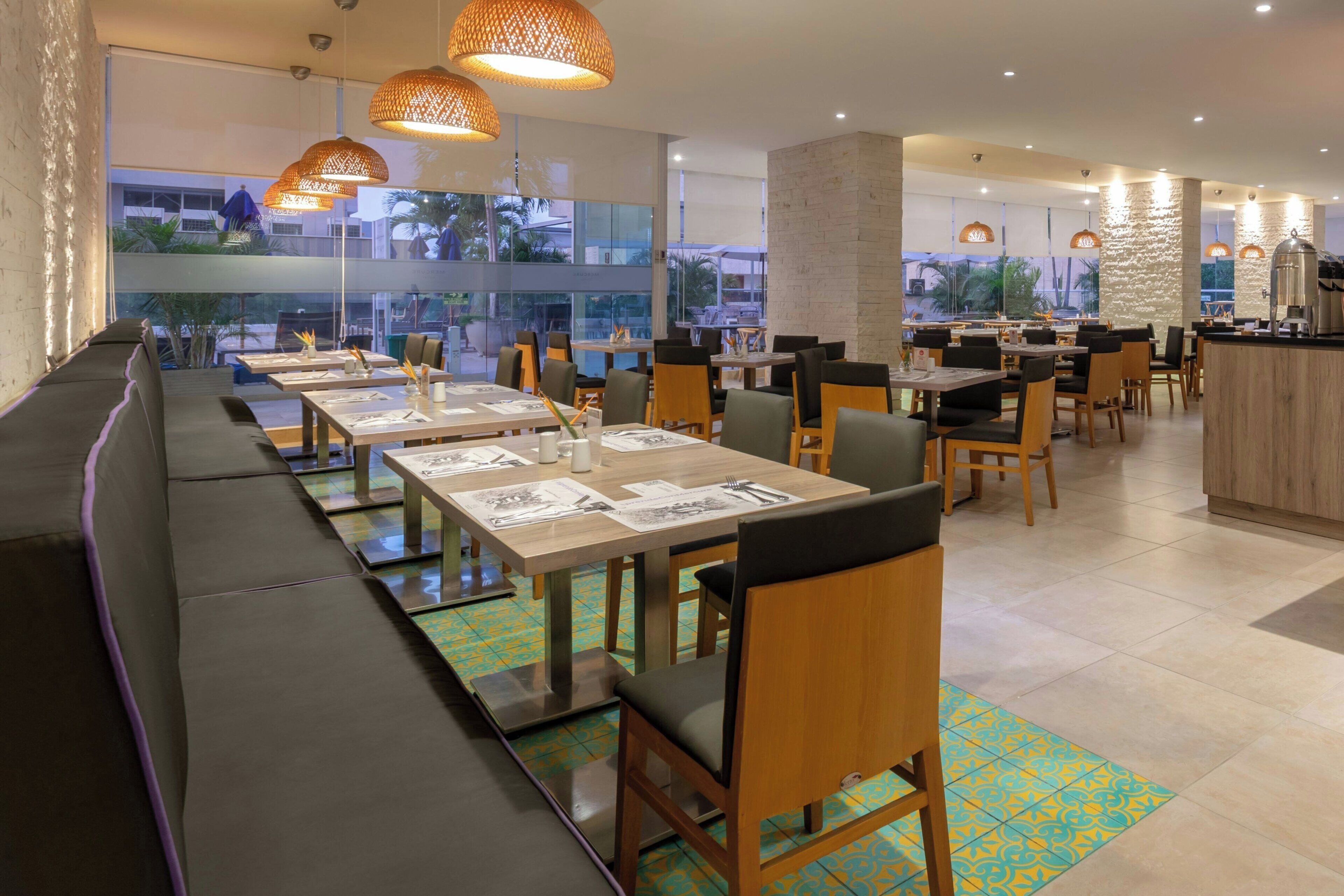 A modern restaurant with long communal tables, cushioned seating, and pendant lighting.