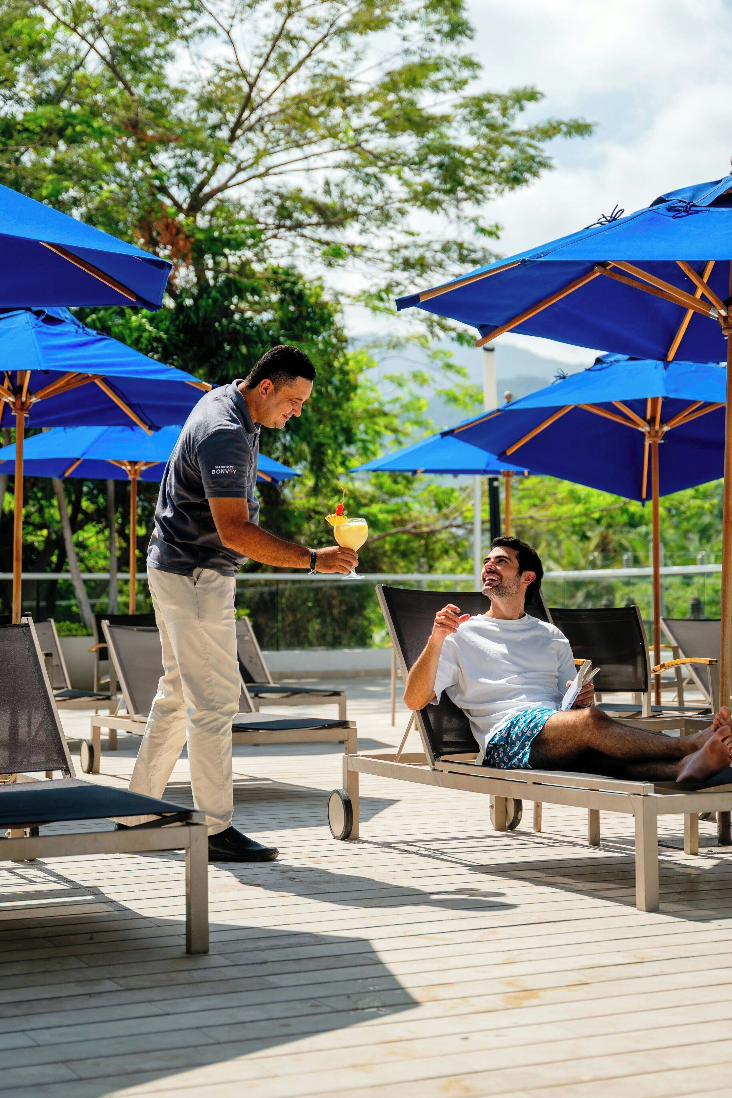 A man serving a drink to a seated man at a poolside area with multiple blue umbrellas and lounge chairs.