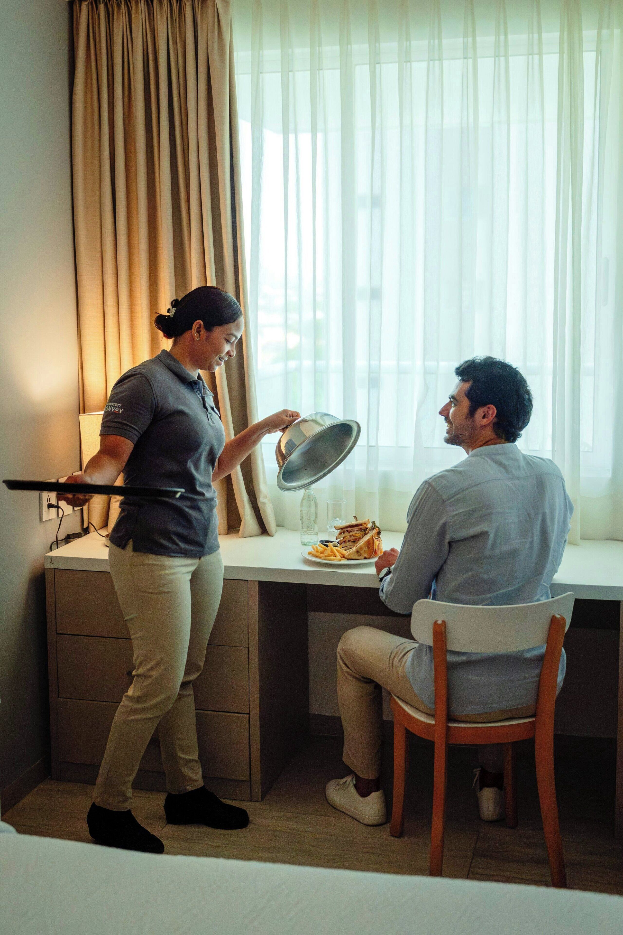A hotel room with a desk, a chair, a dining table with a plate of food, and a window with curtains.