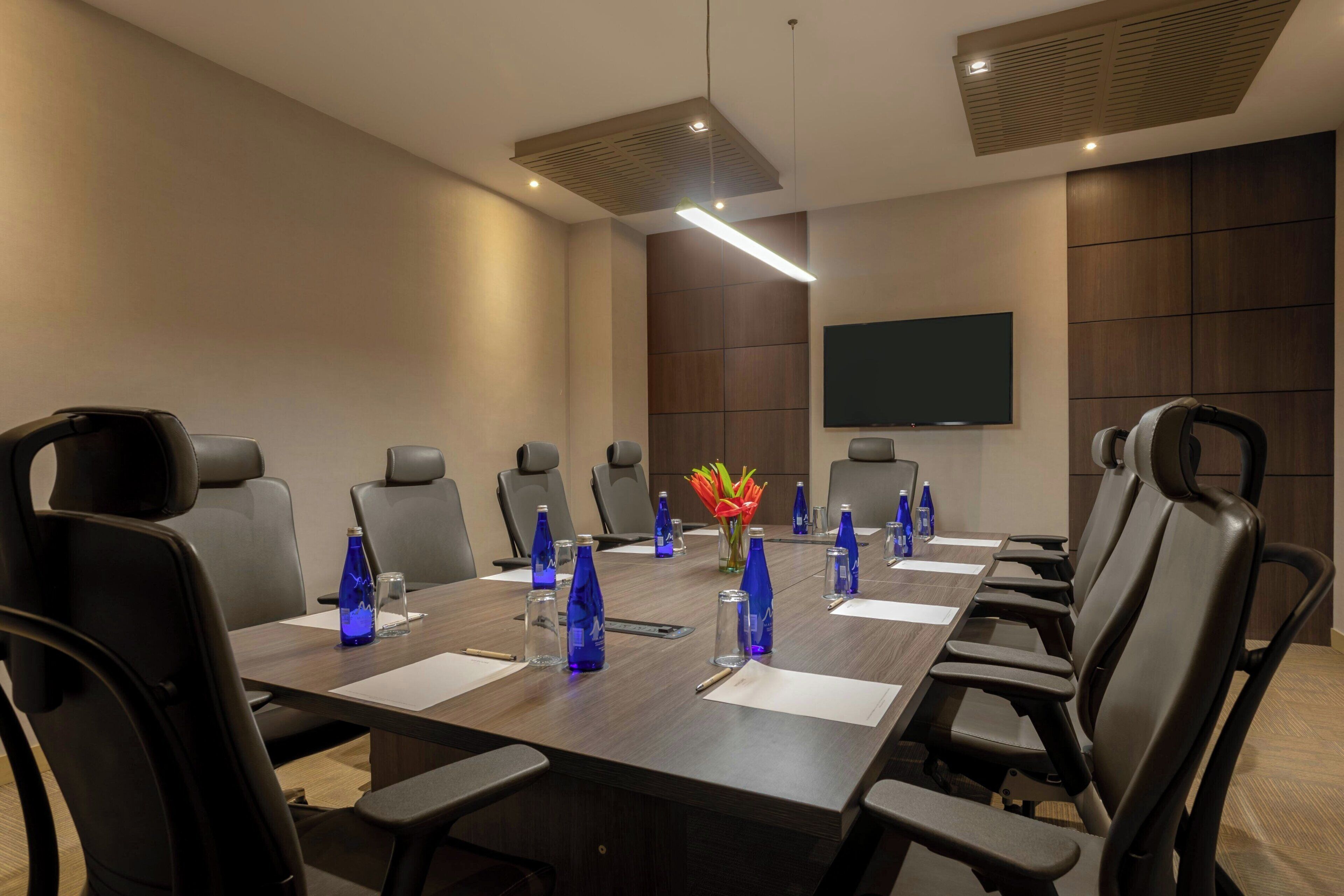 Une salle de conférence avec une longue table, des chaises noires et un téléviseur à écran plat fixé au mur.