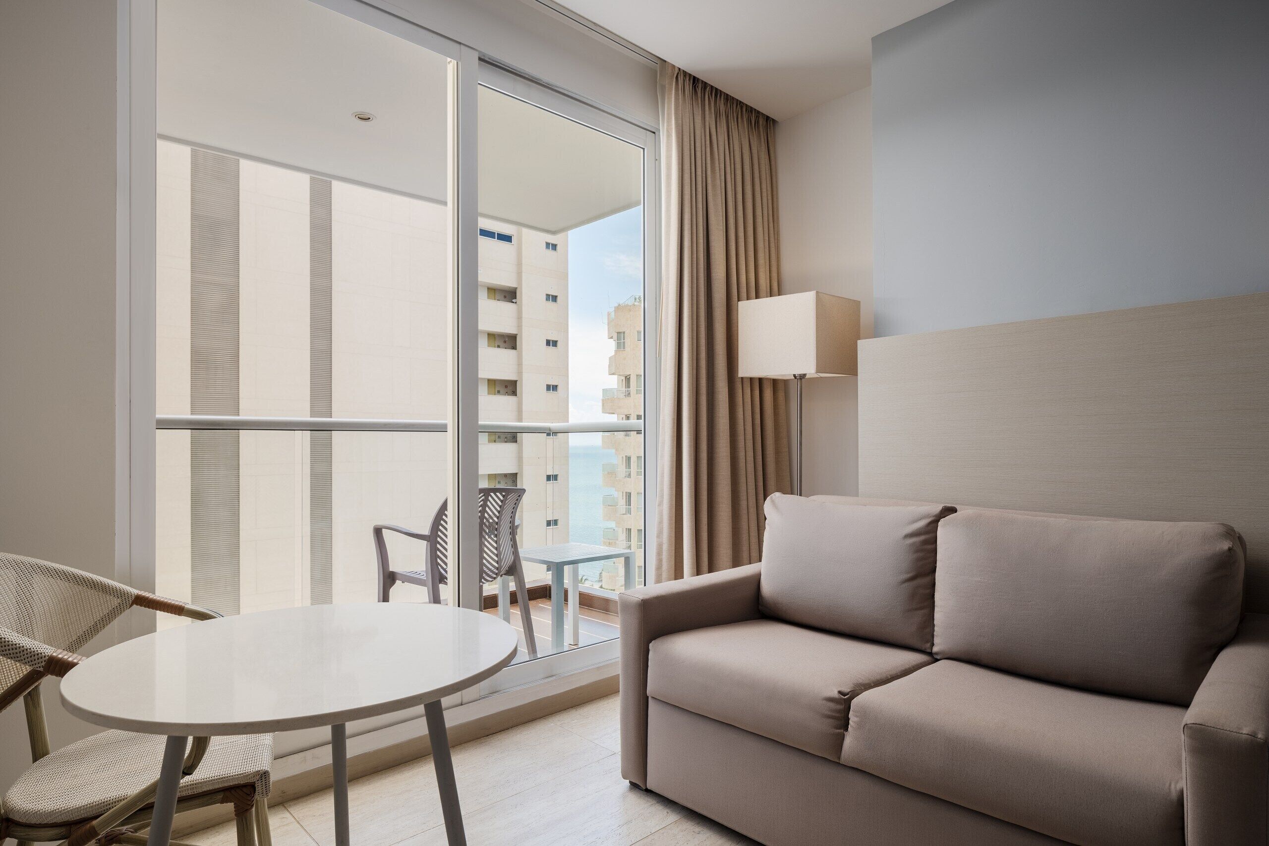 Una habitación de hotel moderna con sofá, una mesita redonda y una silla. Cuenta con un ventanal con vistas a edificios.