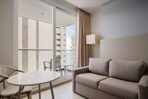 Ein modernes Hotelzimmer mit einer Couch, einem kleinen runden Tisch und einem Sessel. Ein groĂes Fenster bietet Blick auf die Stadt.