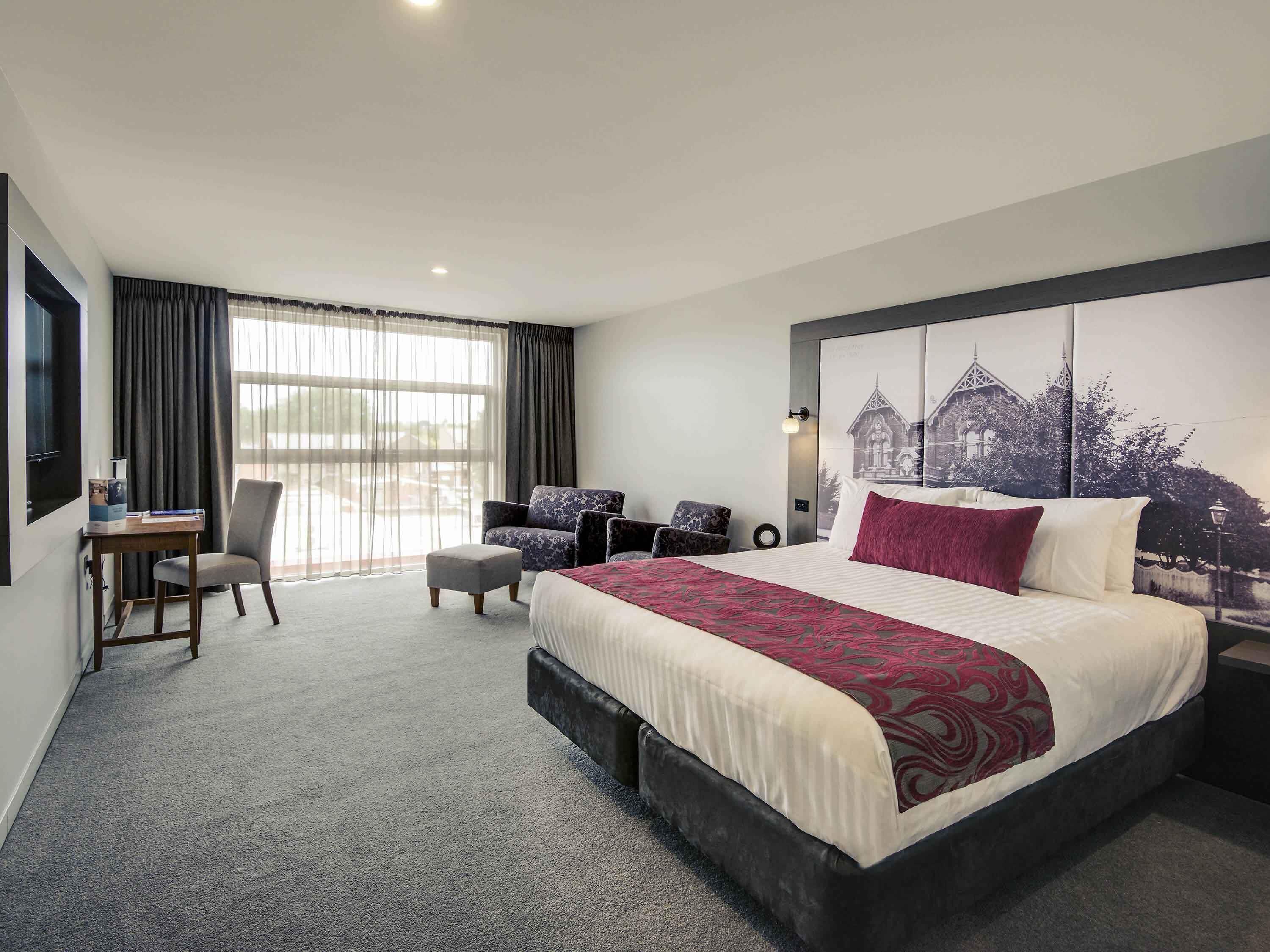 A hotel room with a large bed, a desk, a chair, and a view of a building through the window.