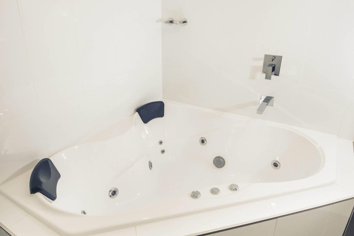 A white bathtub with a built-in seat and multiple jets, a handheld showerhead, and a control panel with buttons and a faucet.
