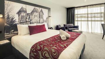A hotel room with a large bed, a headboard with a cityscape photo, a seating area with chairs, and a large window with curtains.